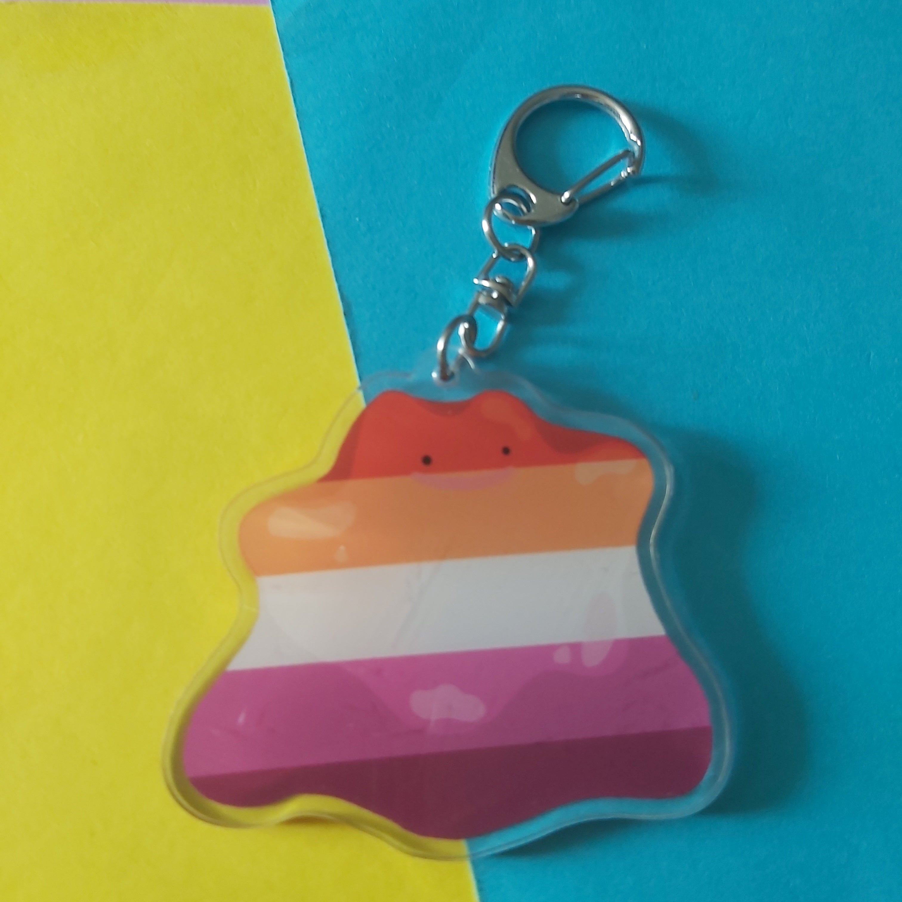 A small acrylic keychain charm with a wavy outline and a silver lobster clasp, featuring horizontal stripes in orange, white, pink, and purple. The charm is photographed on a split yellow and blue background.