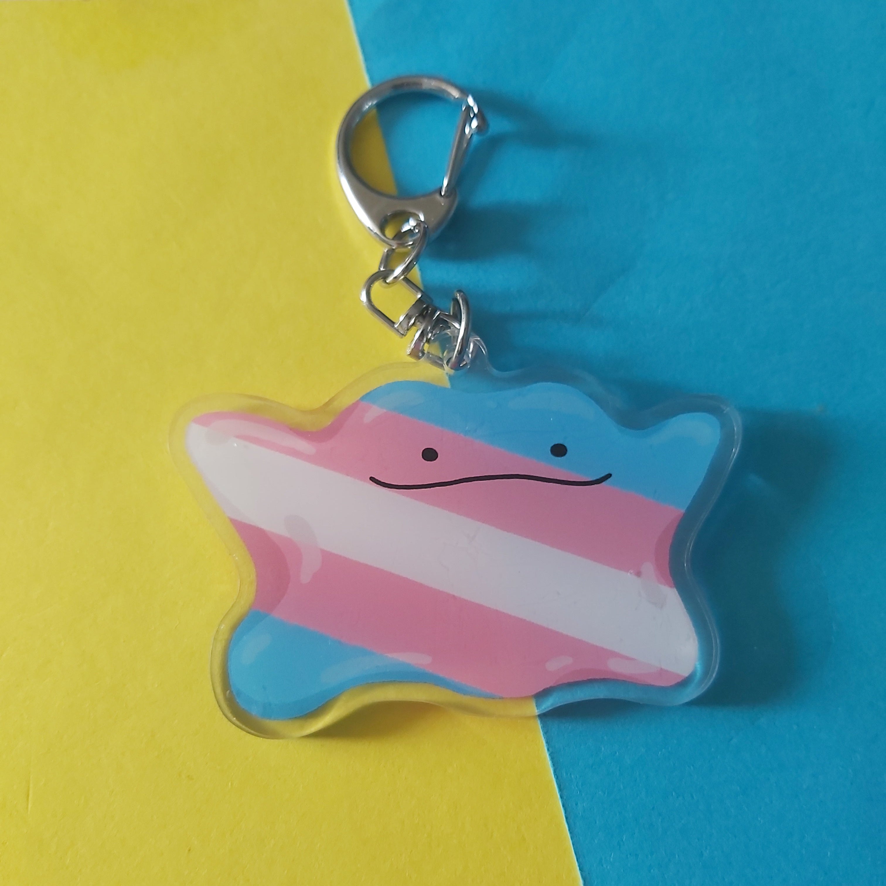 A keychain with a blue, white, and pink striped design and a simple face, attached to a silver metal clasp. The keychain is photographed on a background split diagonally between yellow and blue paper.