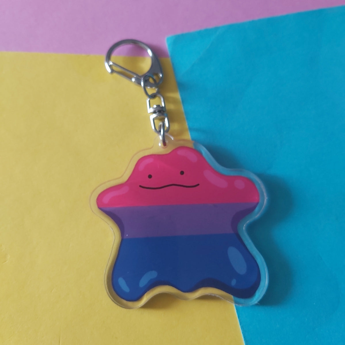 A small acrylic keychain charm shaped like a blob with horizontal pink, purple, and blue stripes, attached to a silver clasp, photographed on a split yellow and blue background with a pink upper section.