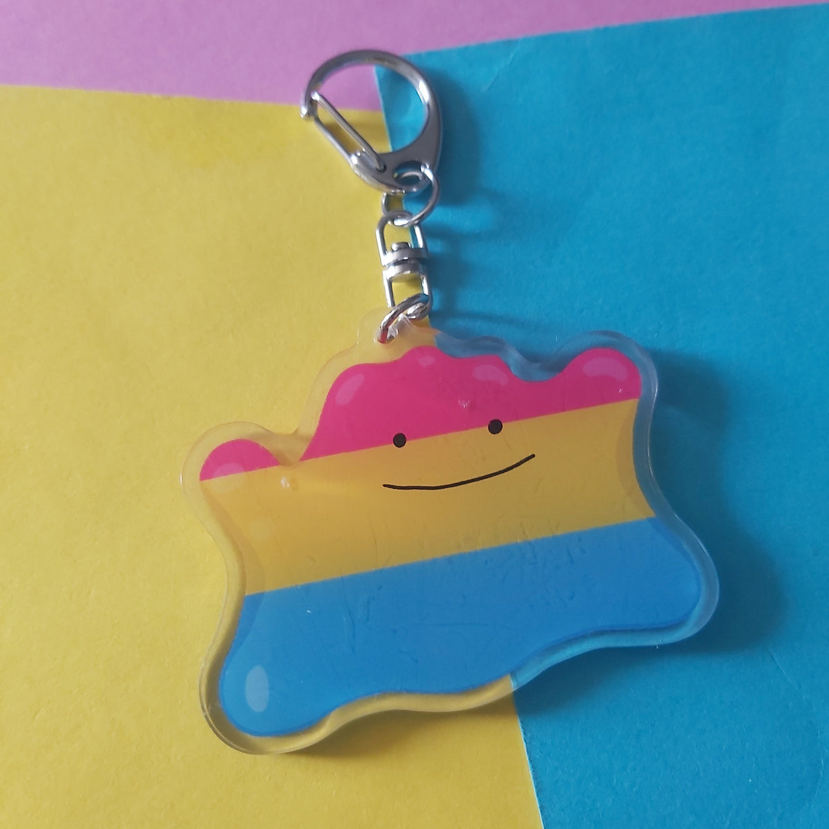 A wavy, cloud-shaped acrylic keychain featuring the pansexual pride flag colours (pink, yellow, and blue) with a simple smiling face, attached to a silver metal lobster clasp. The keychain is photographed on a background split diagonally between yellow and blue paper, with a pink section visible at the top.