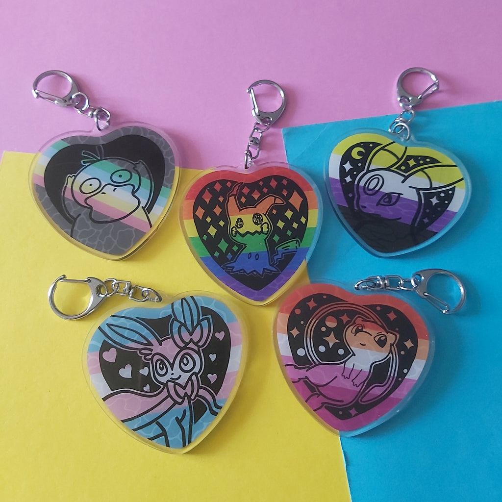 Five heart-shaped acrylic keychains with silver keyrings, each featuring different colorful cartoon character illustrations, arranged on a diagonally split pink, yellow, and blue background.