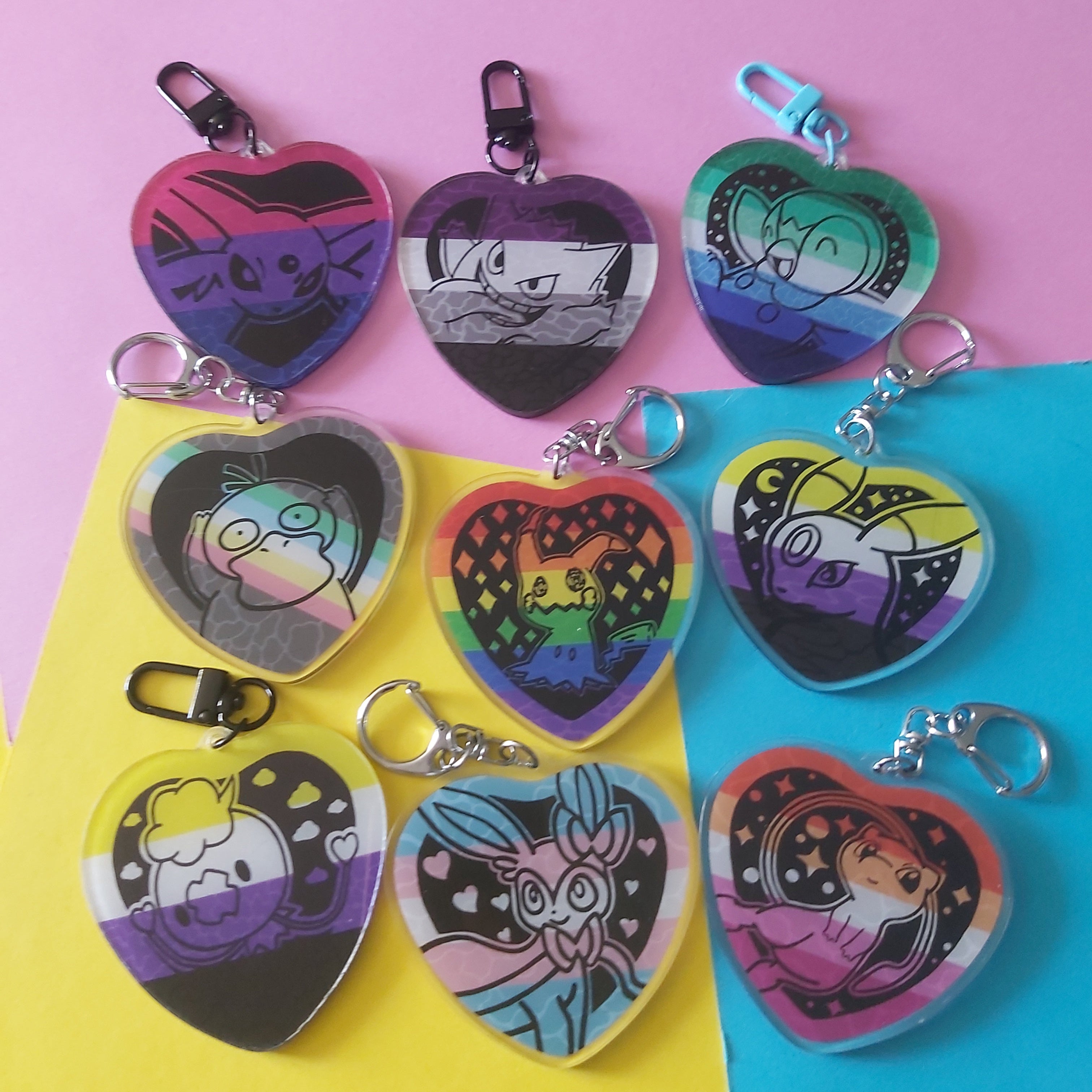 Nine heart-shaped acrylic keychains featuring various Pokémon characters in colourful, stylised designs on Pride flag backgrounds, arranged on a diagonally split pink, yellow, and blue background.