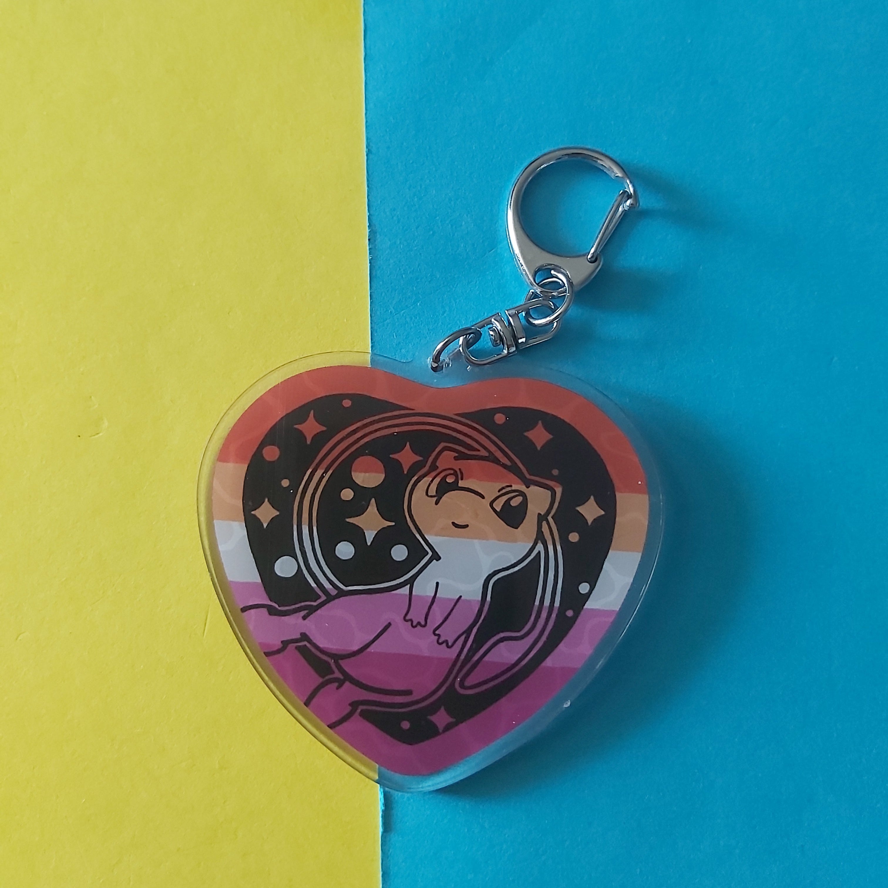 A heart-shaped acrylic keychain featuring the cartoon character Mew on a lesbian pride flag background, attached to a silver metal clasp. The keychain is photographed on a split yellow and blue paper background.