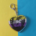 A heart-shaped acrylic keychain featuring an Umbreon Pokémon character illustration with a black, white, yellow, and purple non-binary flag design, attached to a silver lobster clasp, set against a split yellow and blue background.