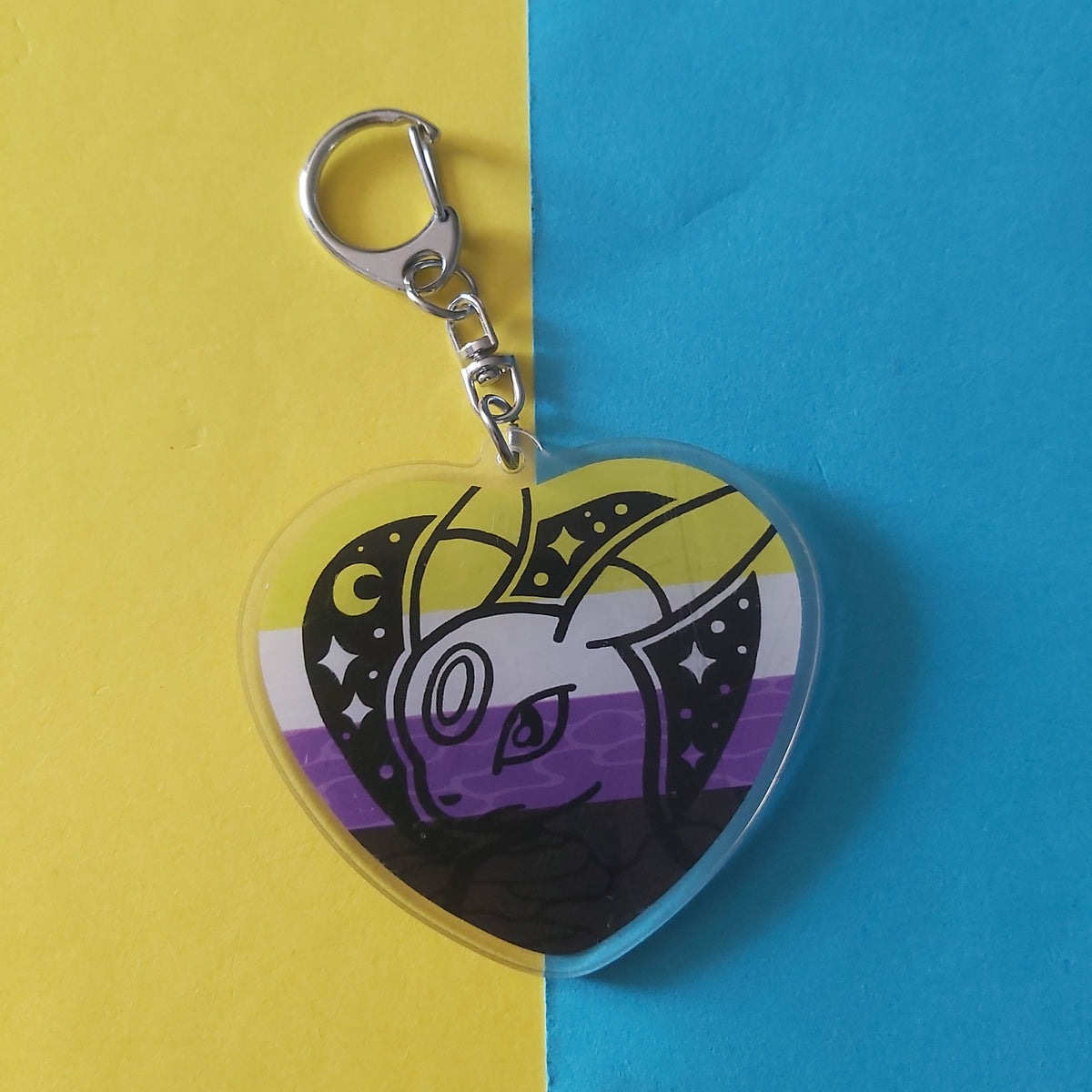 A heart-shaped acrylic keychain featuring an Umbreon Pokémon character illustration with a black, white, yellow, and purple non-binary flag design, attached to a silver lobster clasp, set against a split yellow and blue background.