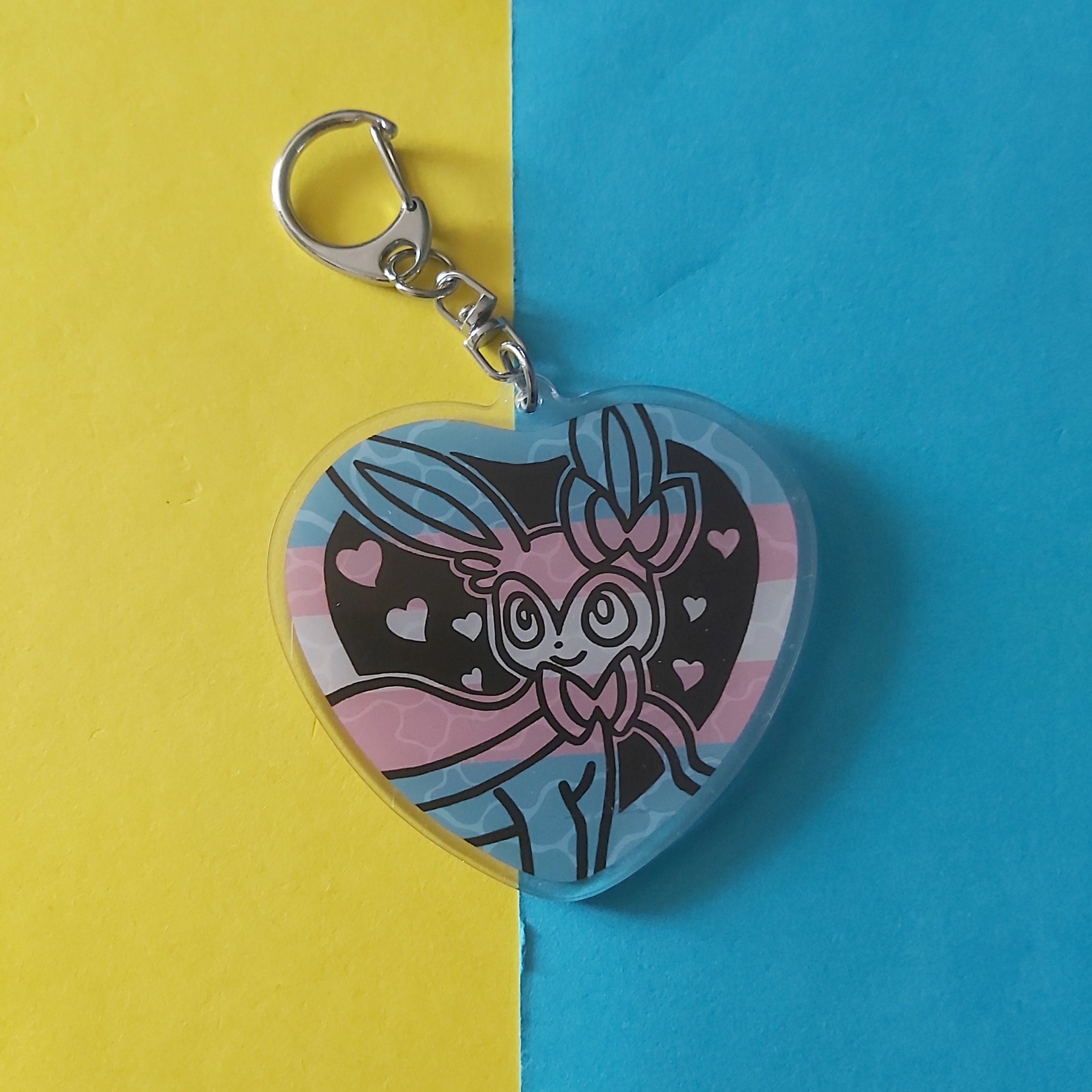 A heart-shaped acrylic keychain featuring a cartoon character Sylveon on a pink, black, and blue striped background, with a silver metal clasp, set against a split yellow and blue paper backdrop.