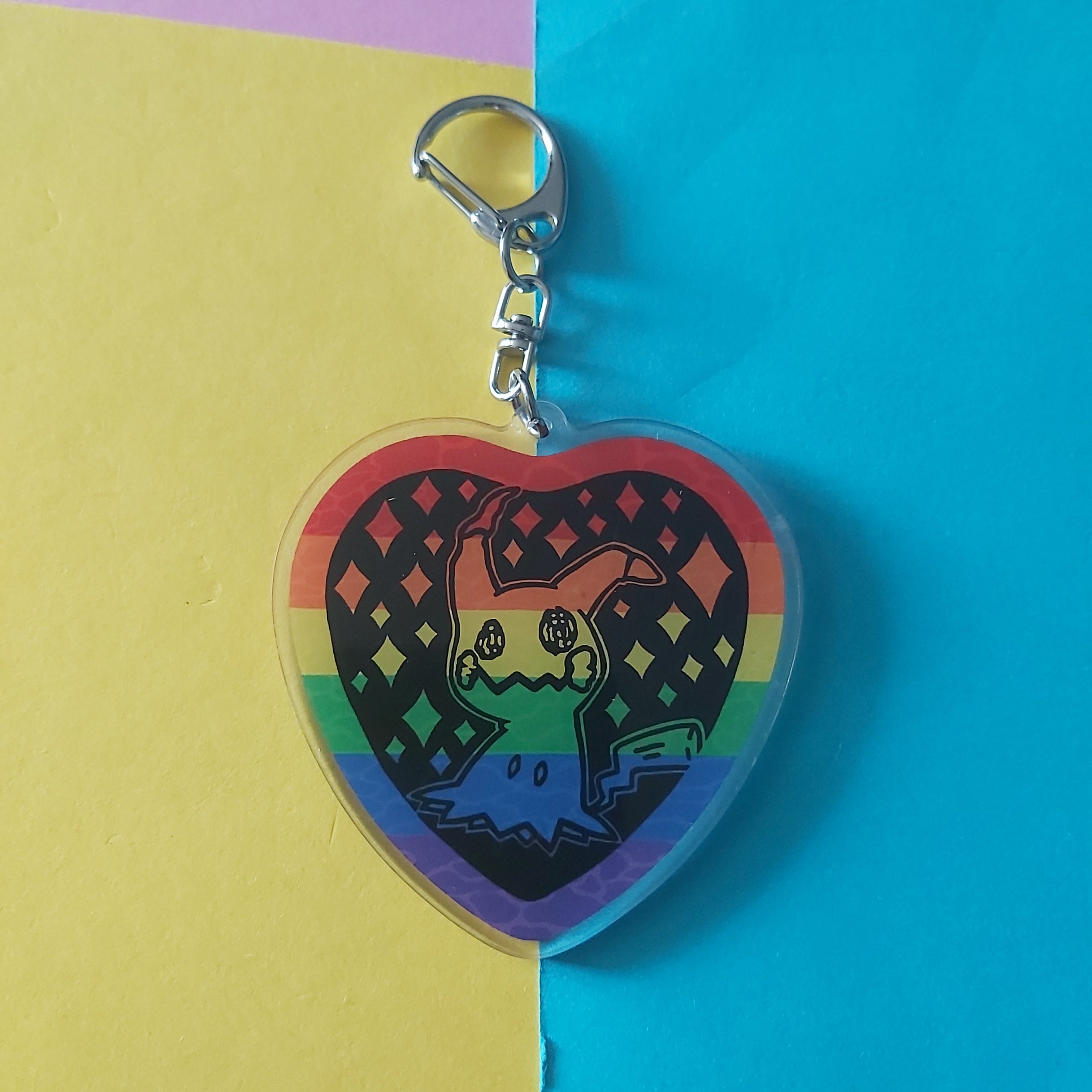 A heart-shaped acrylic keychain featuring a colourful rainbow flag design and a cartoon character Mimikyu, shown on a split yellow and blue background.