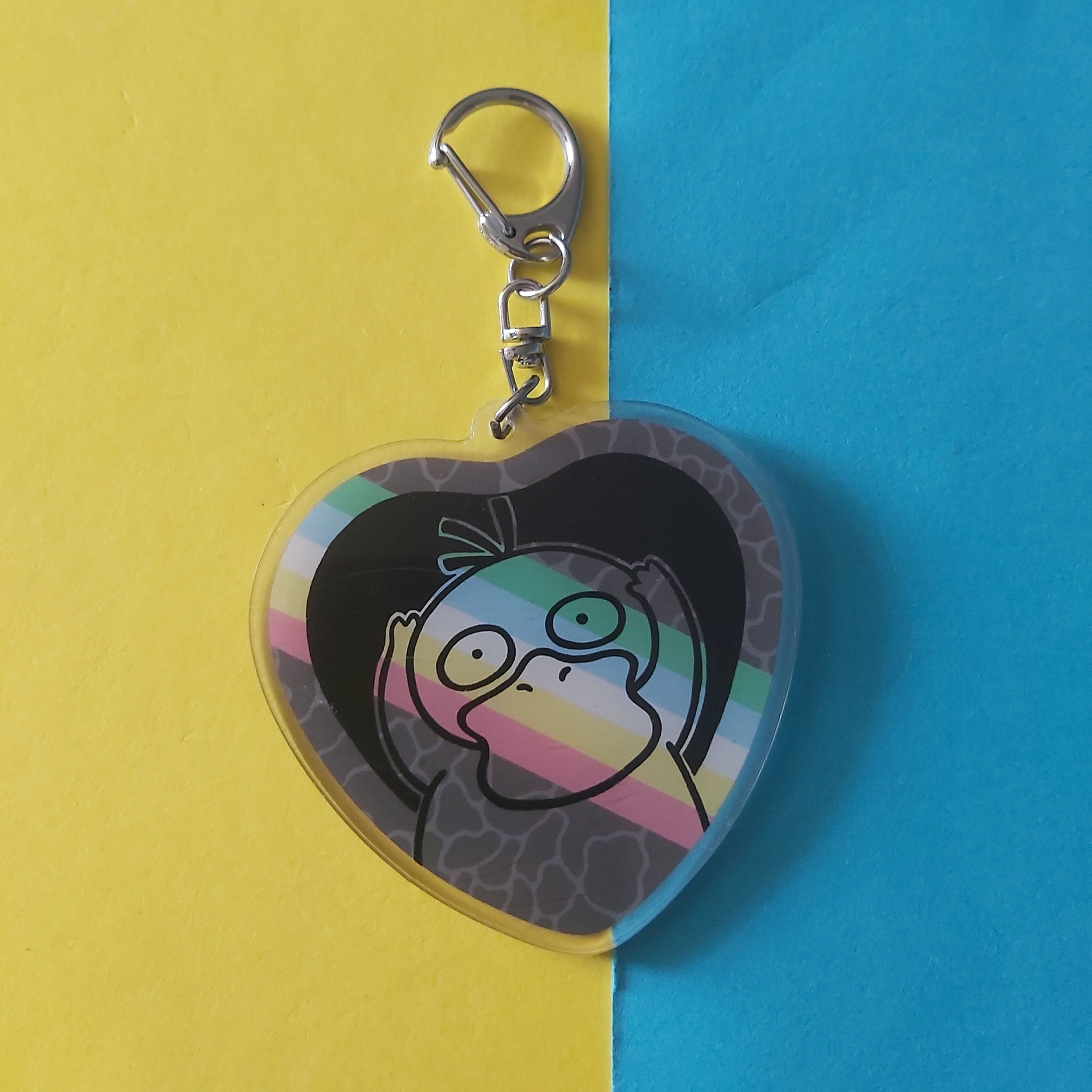 A heart-shaped acrylic keychain featuring a cartoon character, Psyduck, on a disability pride flag, resting on a split yellow and blue background.