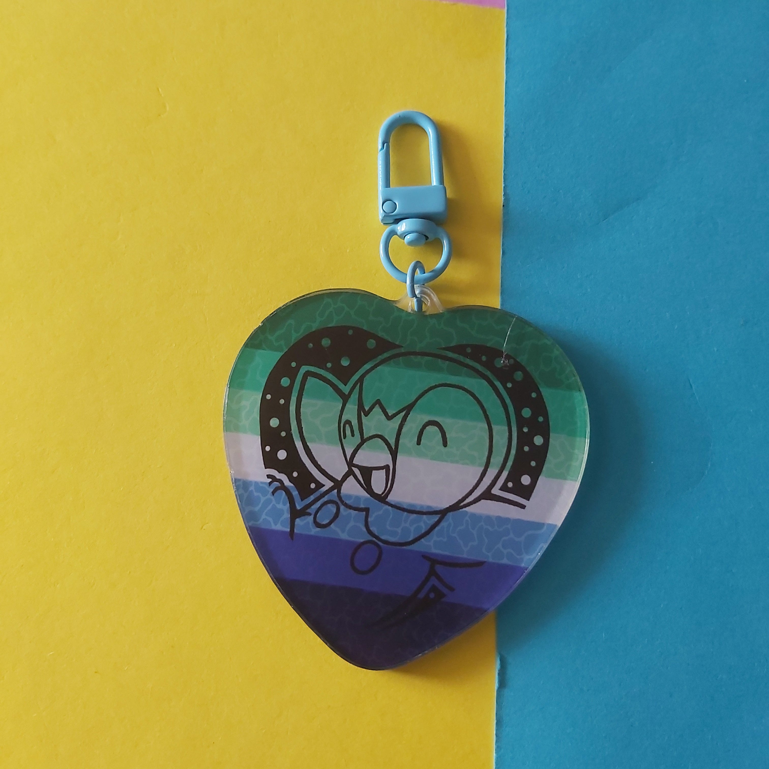 A heart-shaped keychain featuring a cartoon character Piplup on a striped, gay male flag design. The keychain has a light blue clasp and is displayed against a split yellow and blue background.