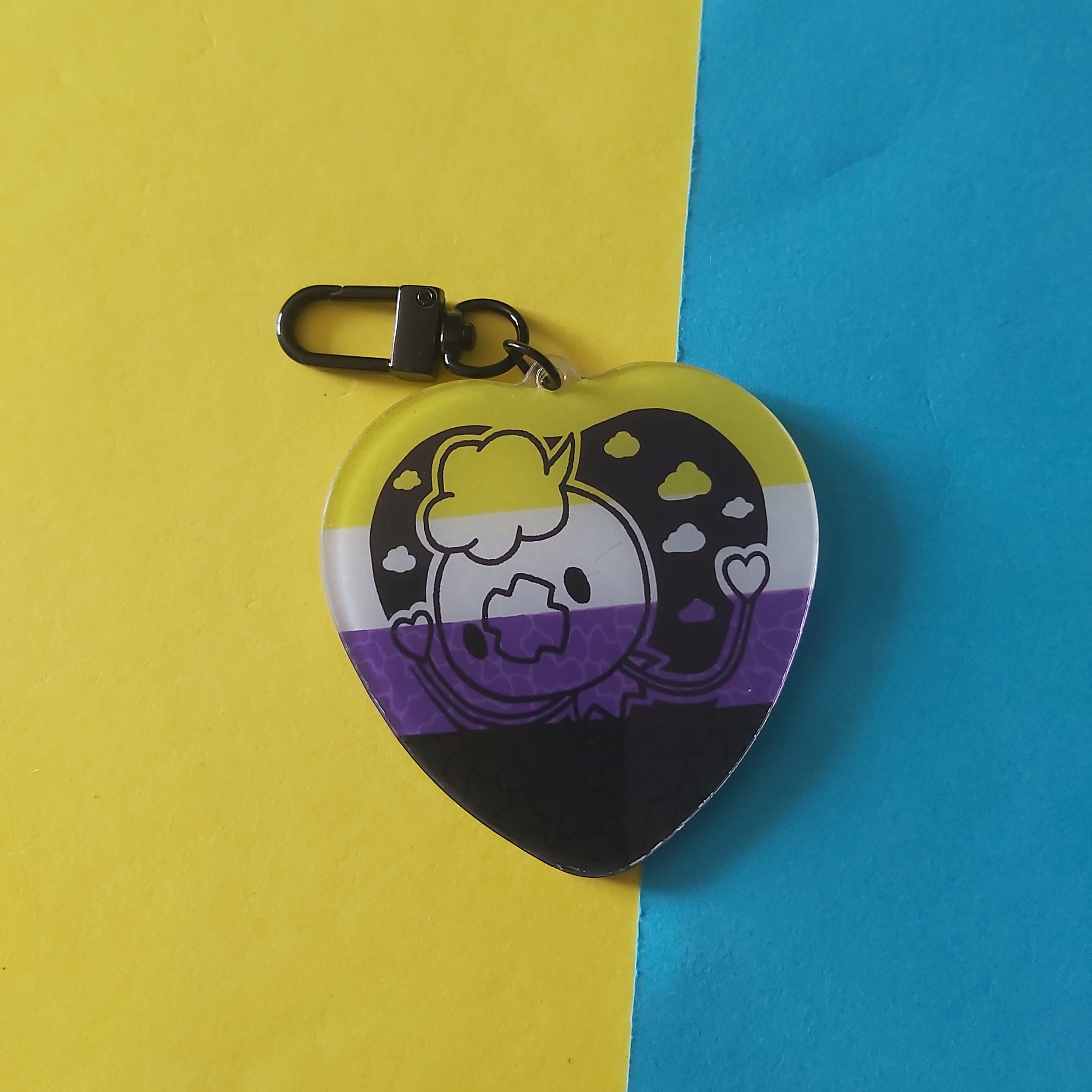 A heart-shaped keychain featuring a cartoon character, Drifloon and the non-binary pride flag colours, displayed on a split yellow and blue background.