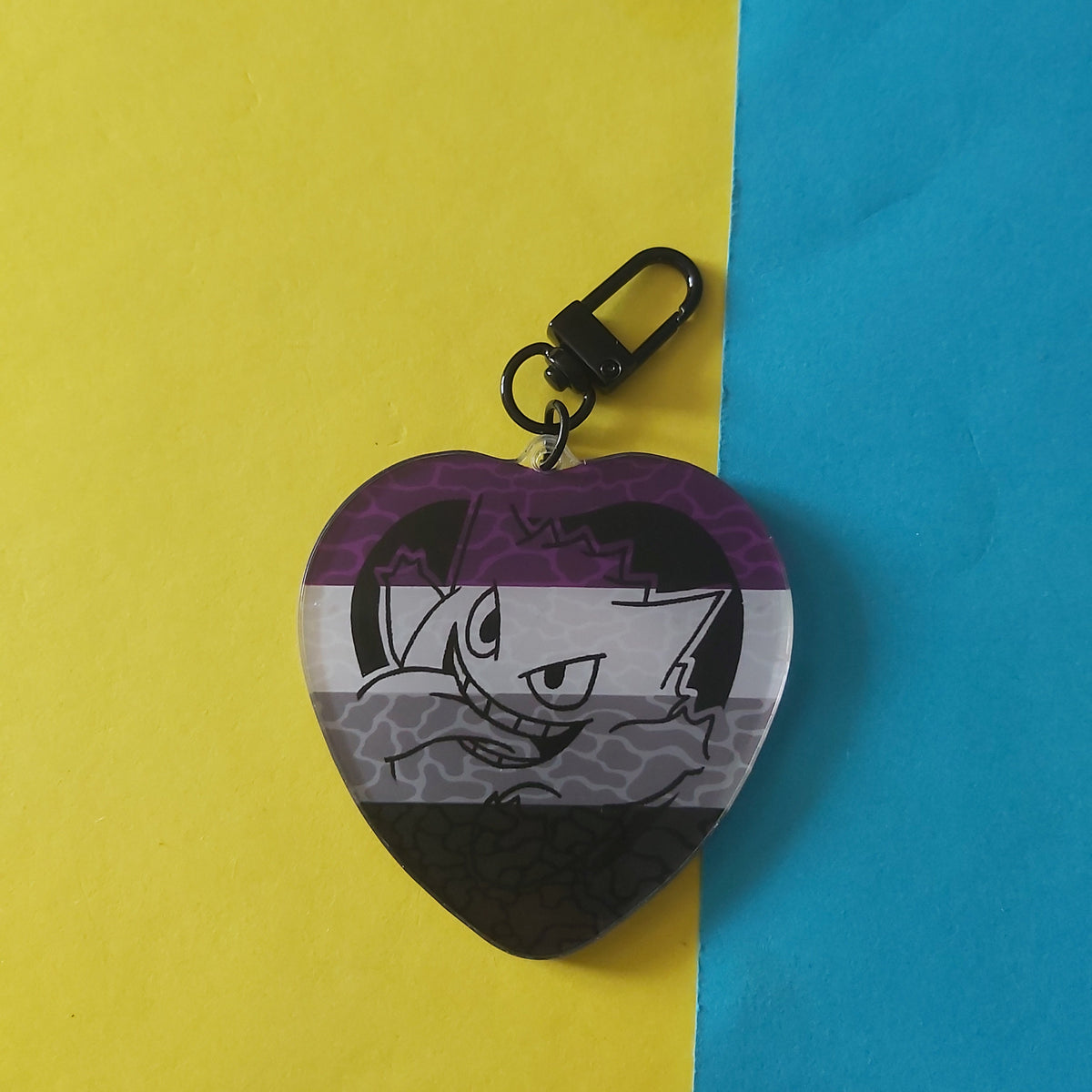 A heart-shaped acrylic keychain featuring the asexual pride flag colours and an illustration of Gengar, attached to a black metal clasp, set against a background of yellow and blue paper.