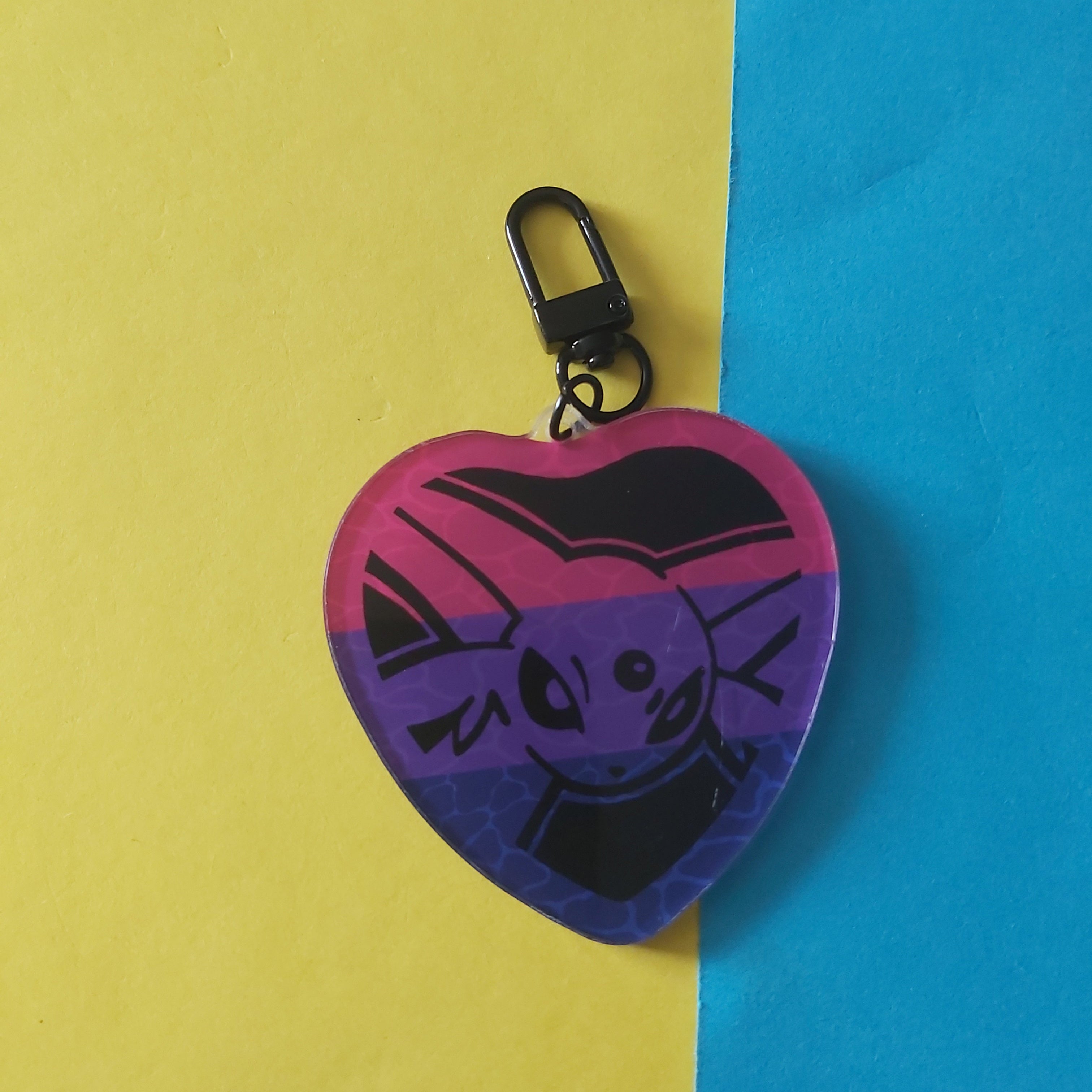 A heart-shaped keychain featuring a stylised cartoon Espeon on a bisexual flag background, attached to a black metal clasp, set against a background split diagonally between yellow and blue paper.