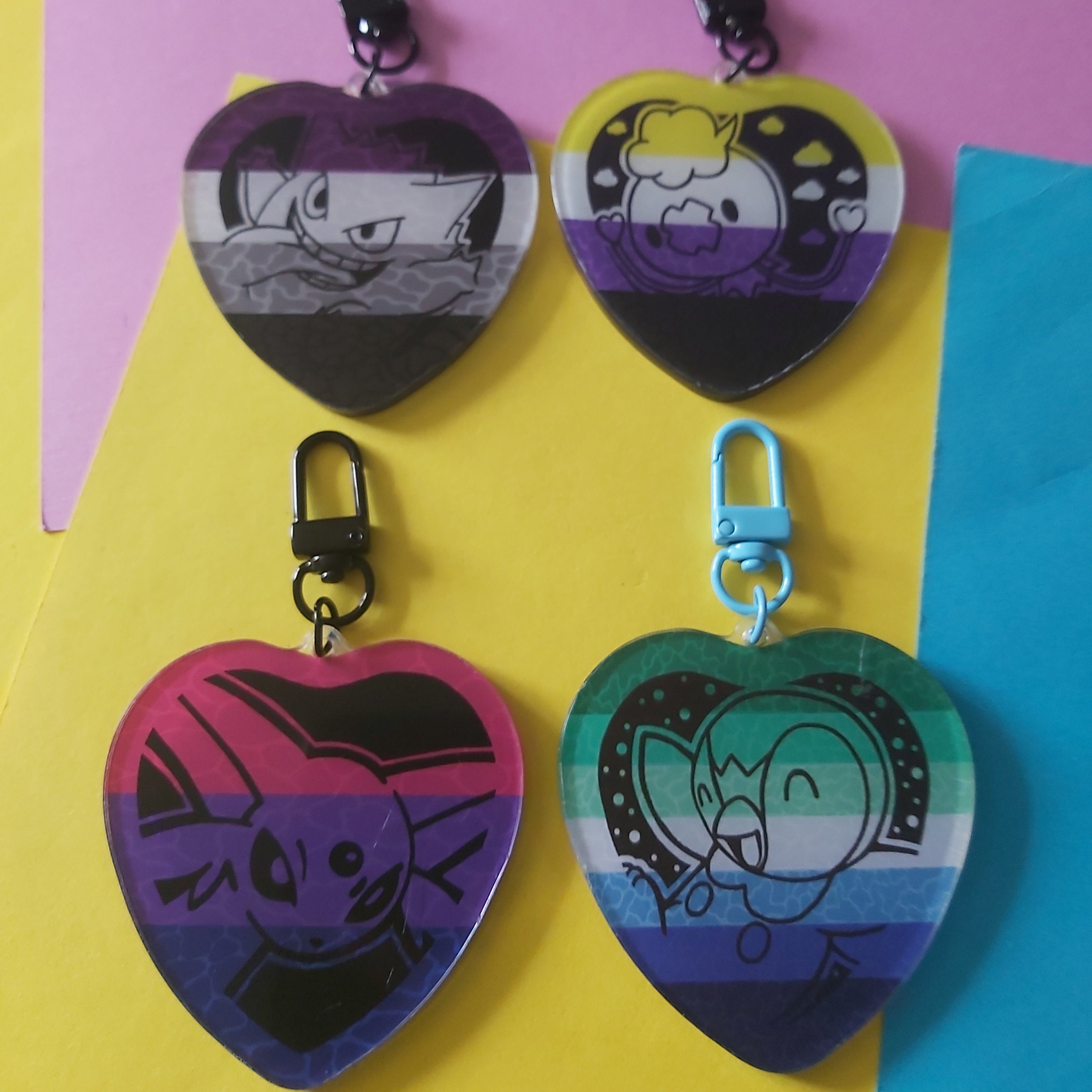 Four heart-shaped keychains with black carabiners, each featuring a different pride flag design and cartoon character illustration, arranged on a colourful background of pink, yellow, and blue paper.