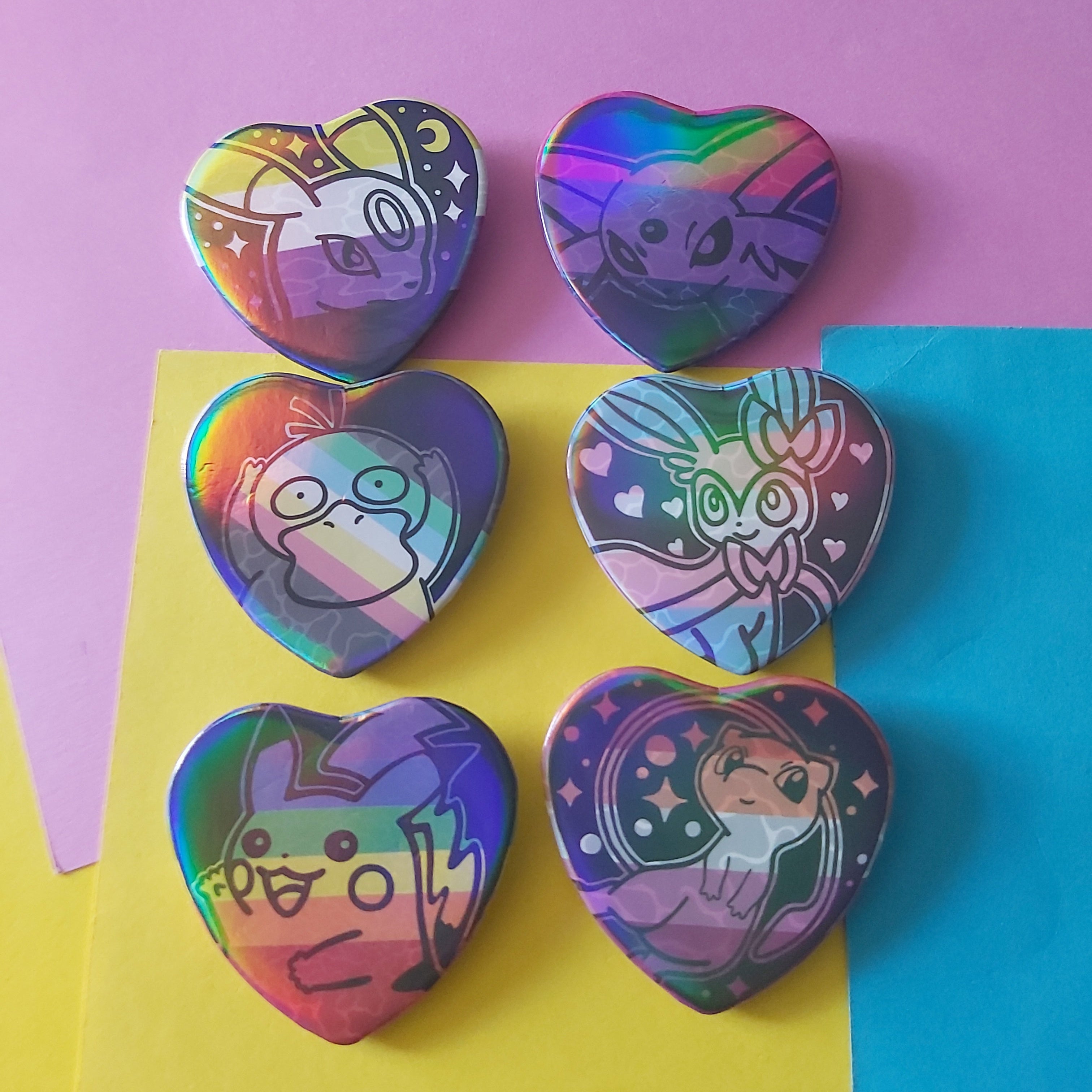 Six heart-shaped holographic pins featuring cartoon characters, arranged on a background of pink, yellow, and blue paper.