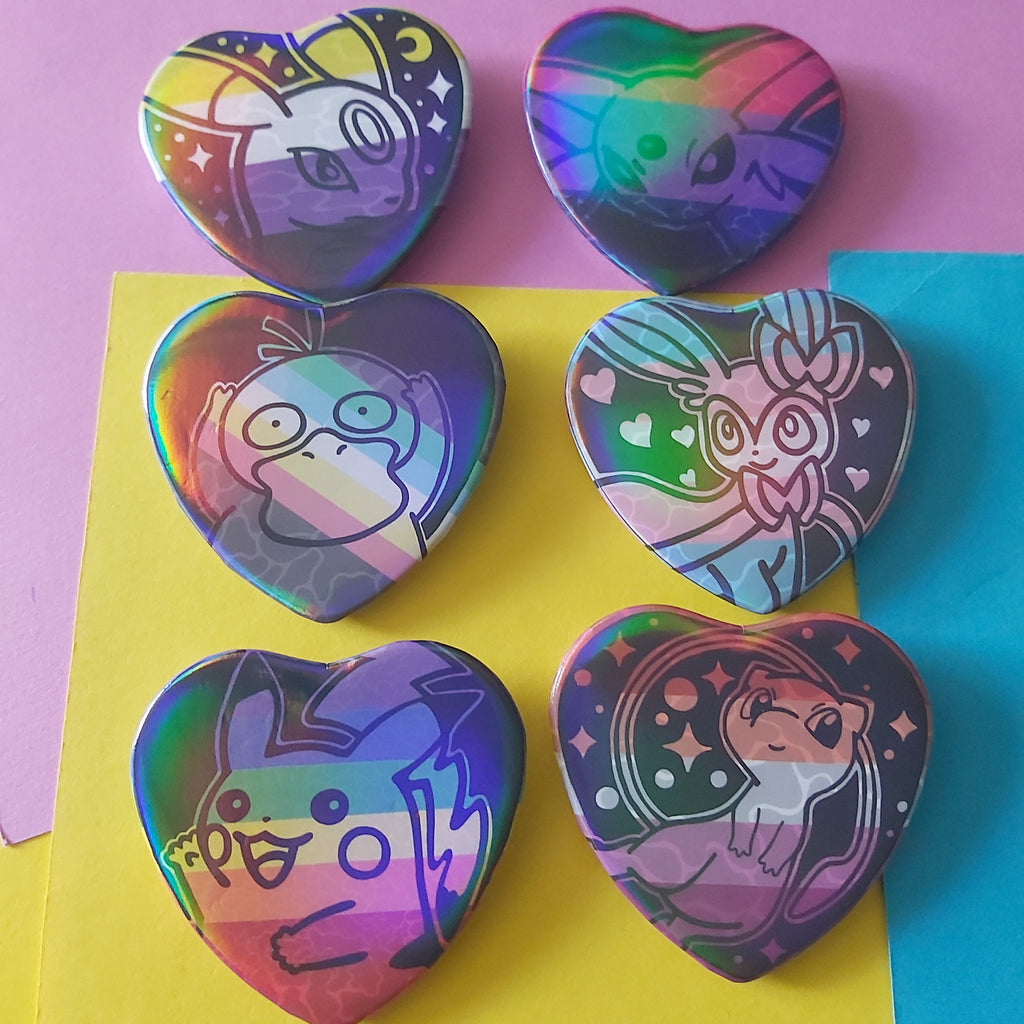 Six heart-shaped holographic pinback buttons featuring cartoon characters, arranged on a background of pink, yellow, and blue paper.