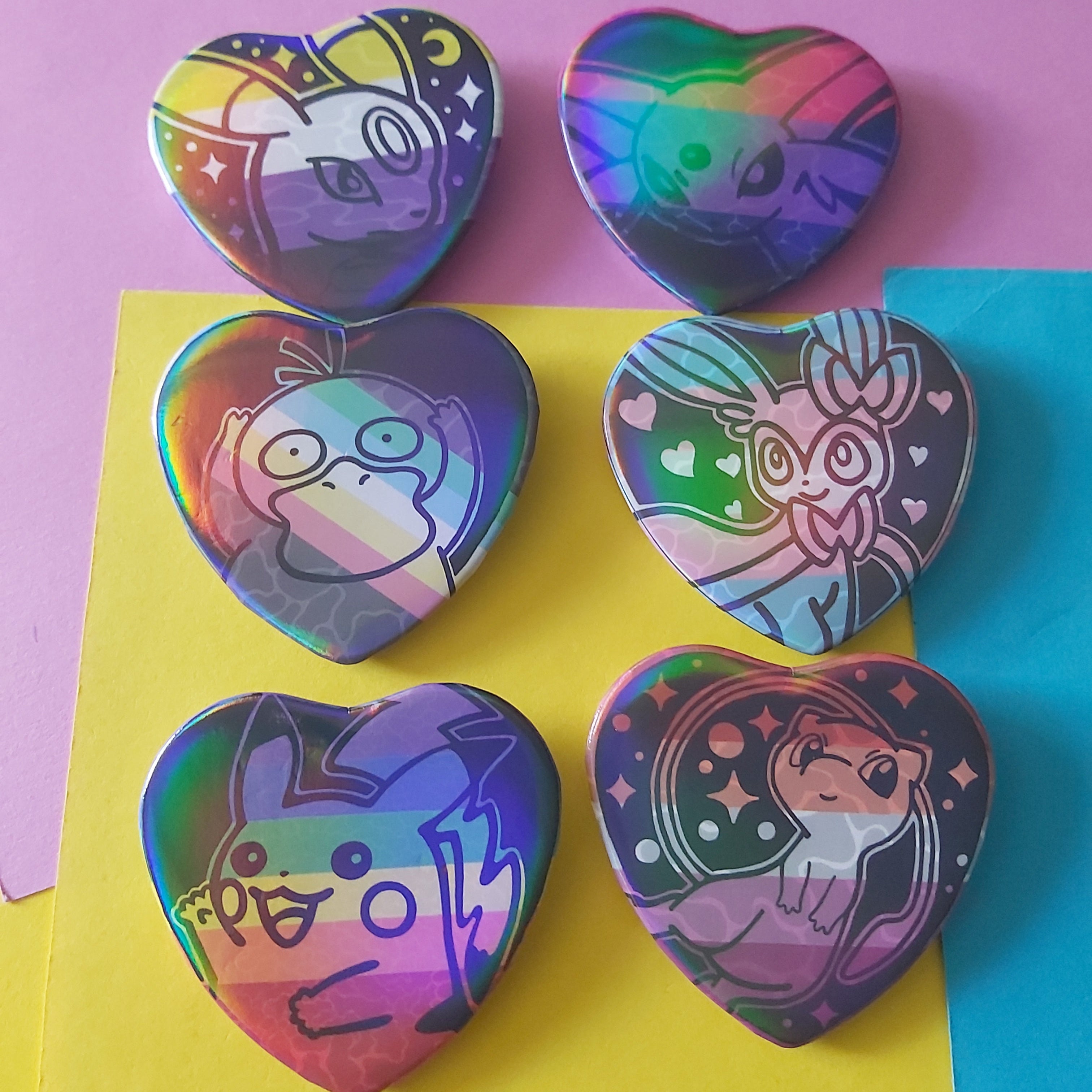 Six heart-shaped holographic pinback buttons featuring cartoon characters, arranged on a background of pink, yellow, and blue paper.