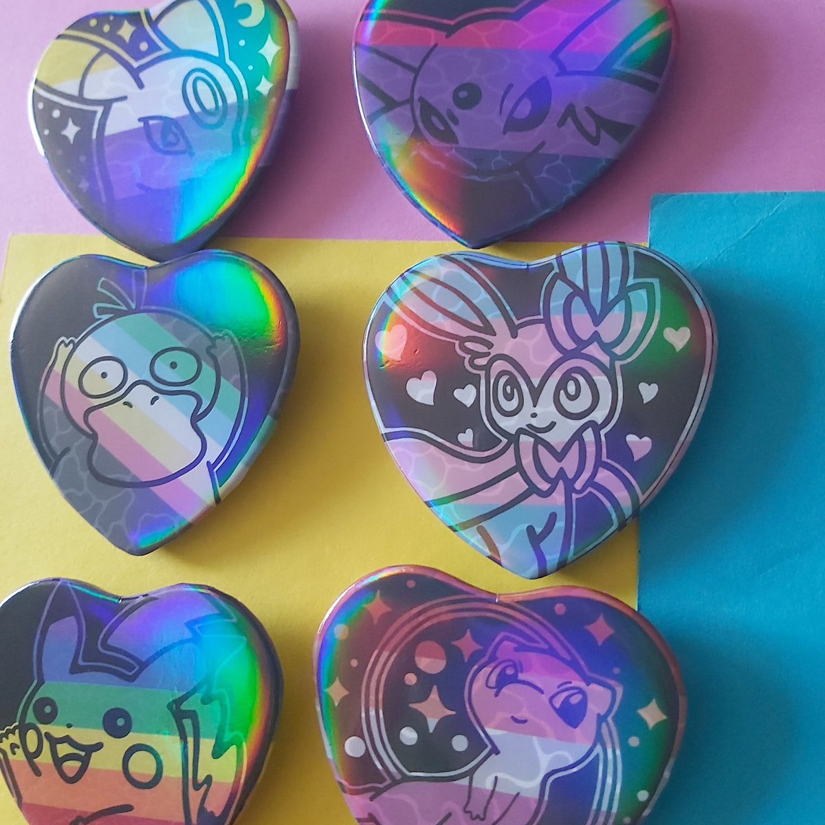Six heart-shaped holographic pins featuring cute cartoon characters, arranged on a colourful background of pink, yellow, and blue paper.
