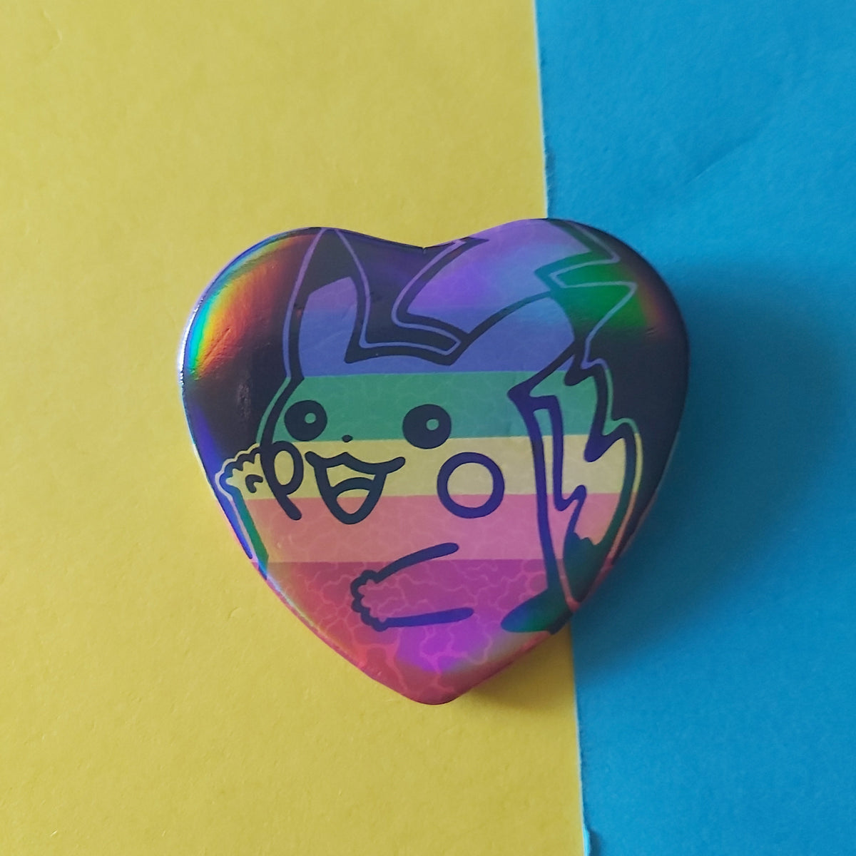 A heart-shaped pin badge featuring a cartoon character with a rainbow flag pattern, set against a split yellow and blue background.