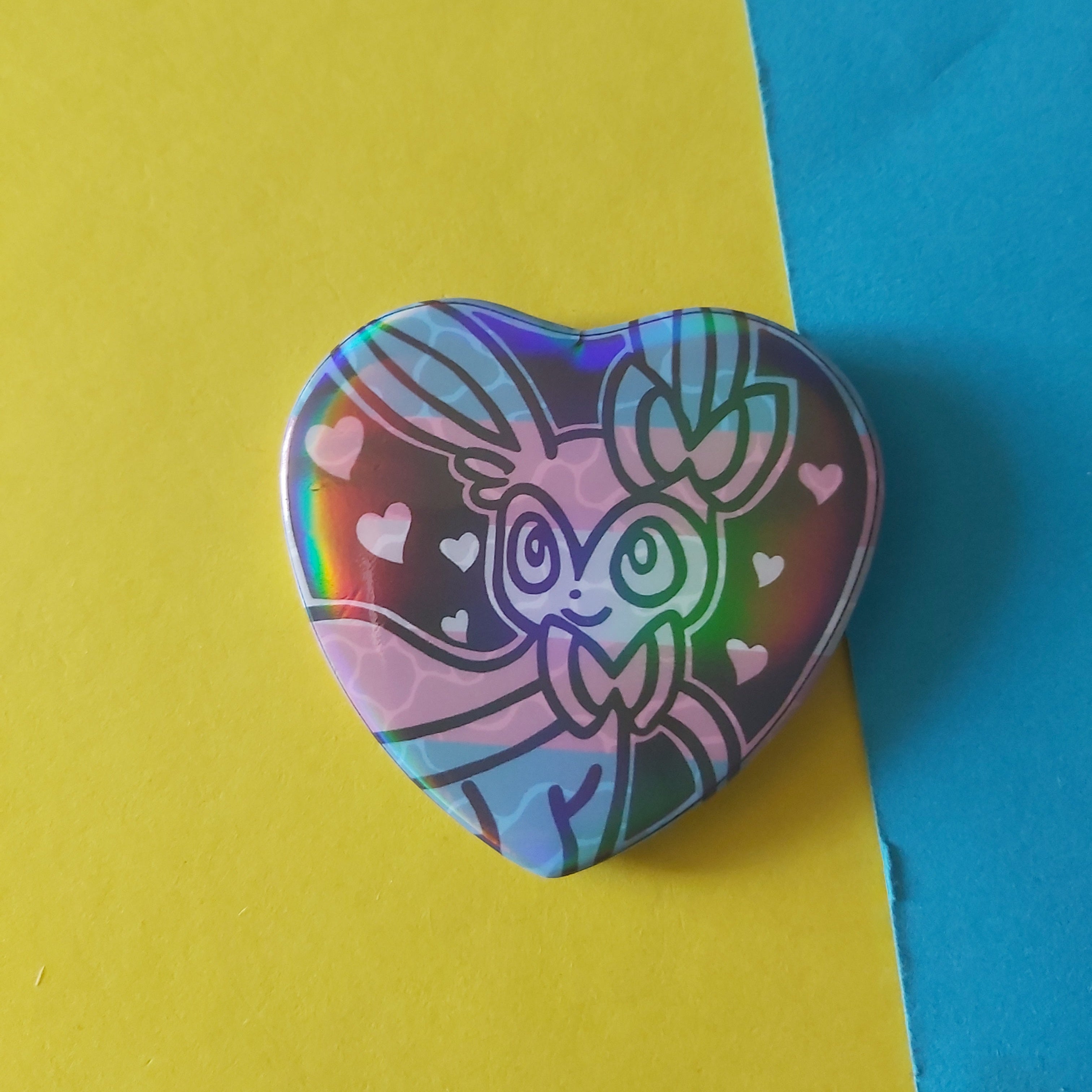 A heart-shaped pinback badge featuring a holographic, iridescent design of the Pokémon Sylveon with pink hearts on a trans pride flag, displayed on a split yellow and blue background.