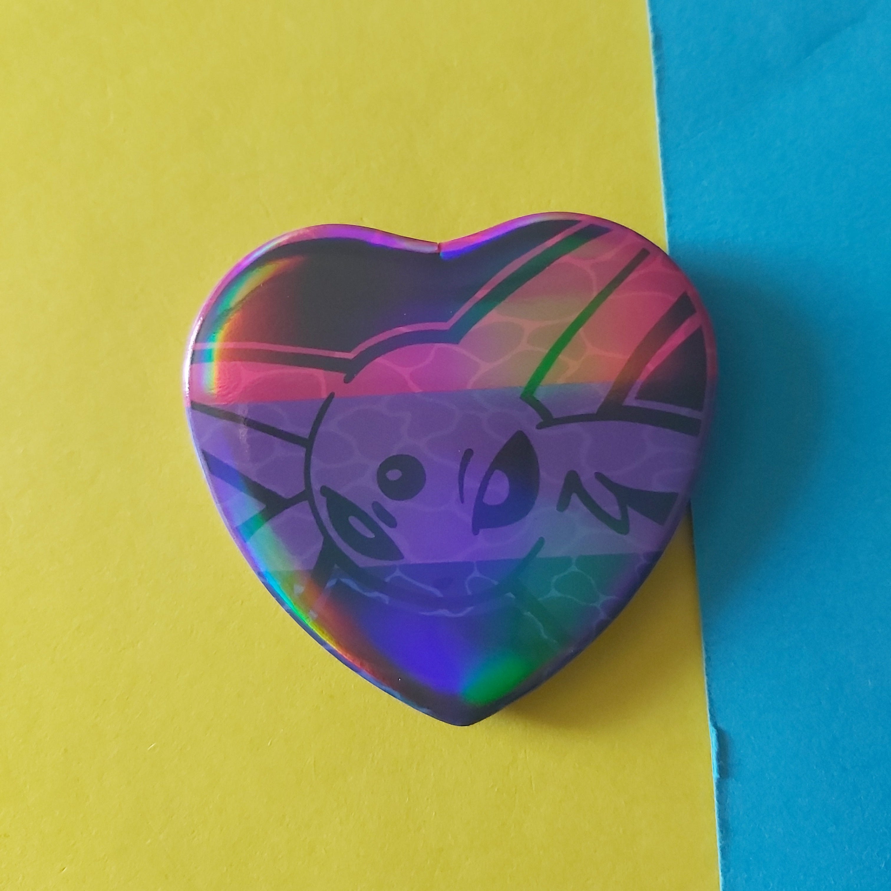 A heart-shaped holographic pinback button featuring a stylised cartoon face and bi flag background, set against a split yellow and blue backdrop.