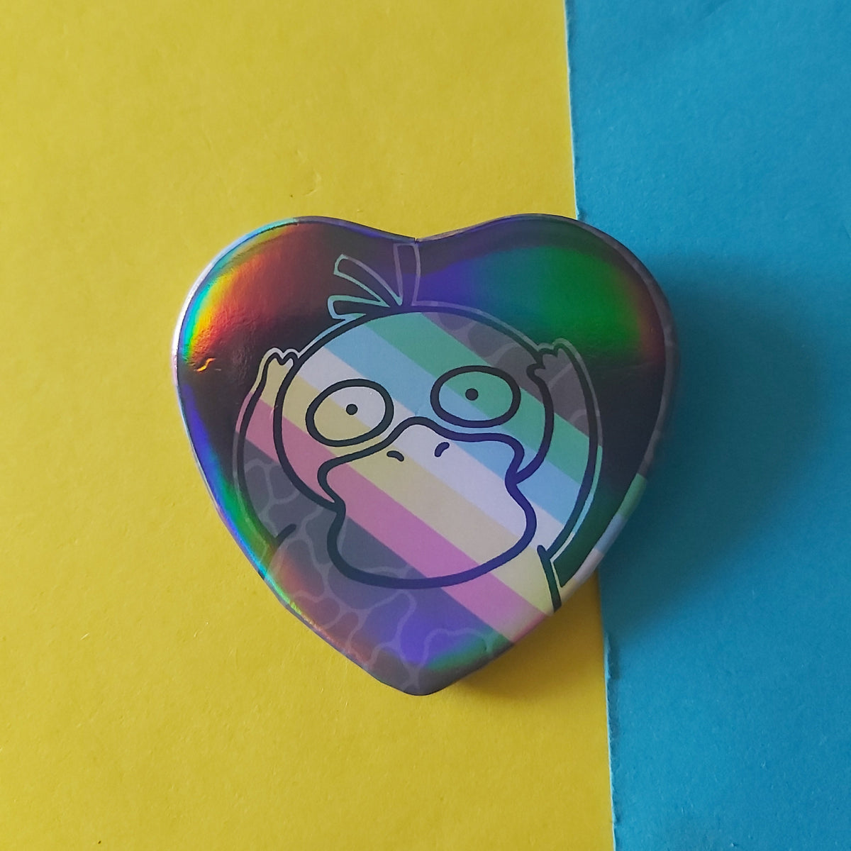 A heart-shaped holographic pinback button featuring a cartoon character on the disability pride flag, set against a split yellow and blue background.