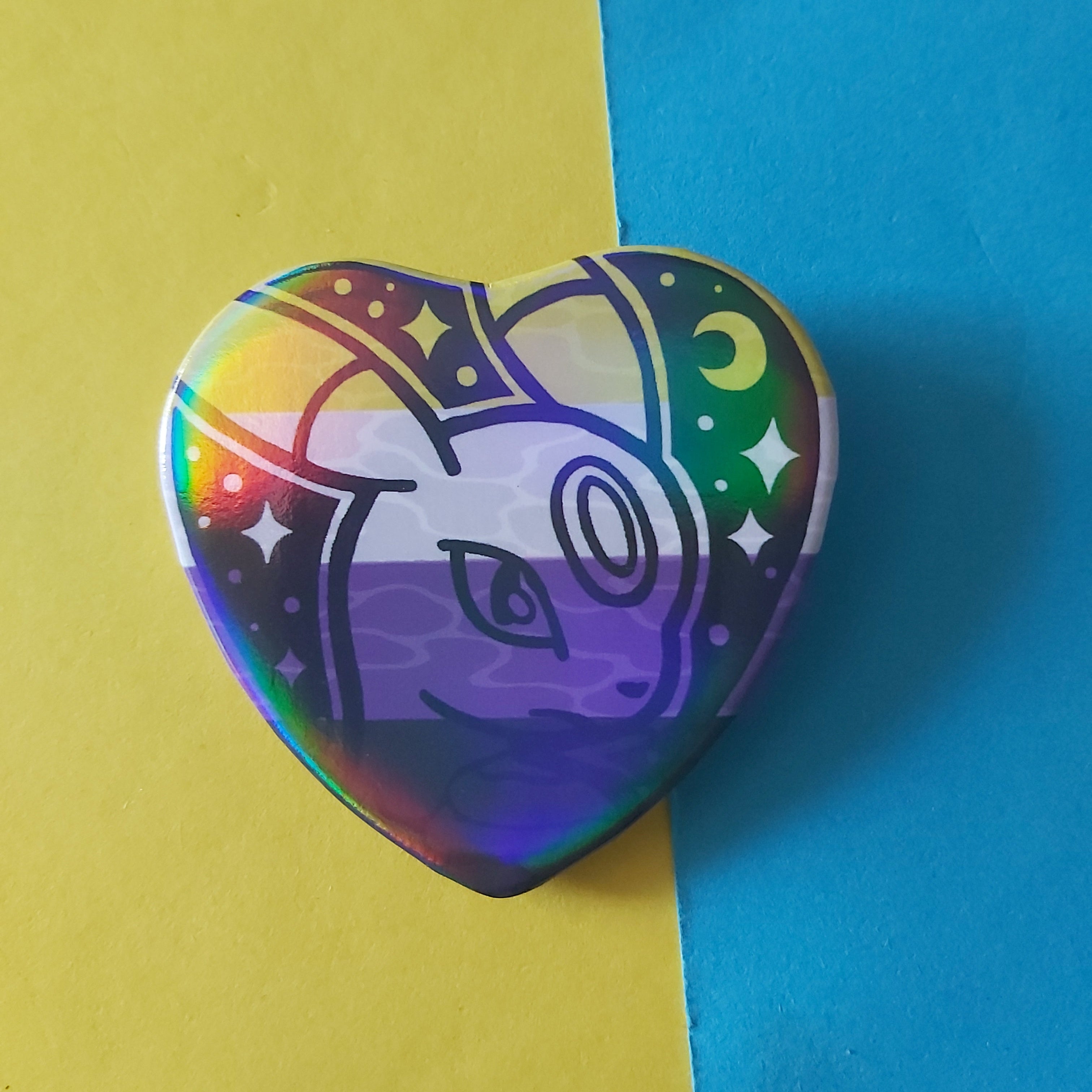 A heart-shaped pin featuring a stylized umbreon on a non binary flag background with rainbow-colored holographic finish, photographed on a split yellow and blue background.