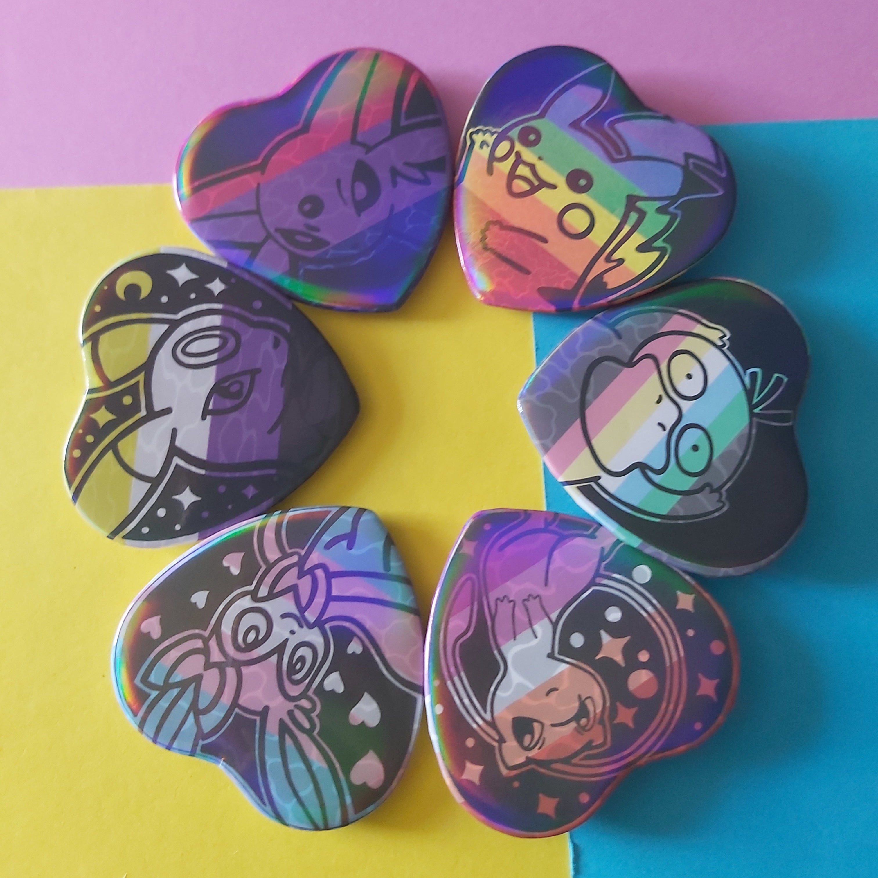 Six heart-shaped badges arranged in a circle on a split yellow and blue background, featuring colourful cartoon character designs and holographic rainbow reflections.