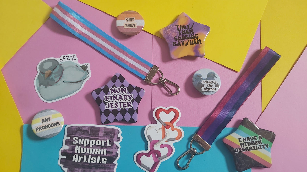 A flat-lay arrangement of various LGBTQ+ pride-themed stickers, pins, and keychains on a colorful geometric background of pink, yellow, and blue paper.