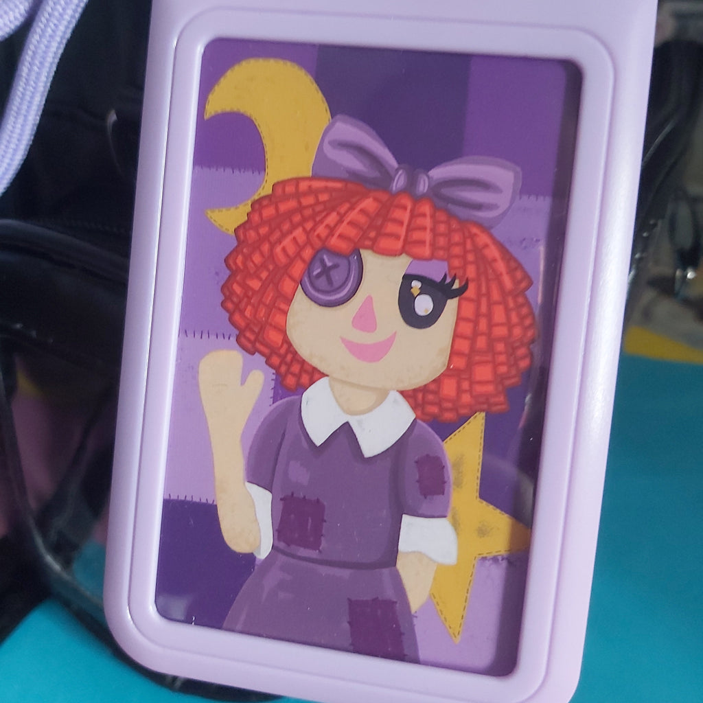 A lavender plastic ID badge holder featuring a cartoon illustration of a girl with red hair, a purple bow, and a purple dress, shown resting against a black handbag.