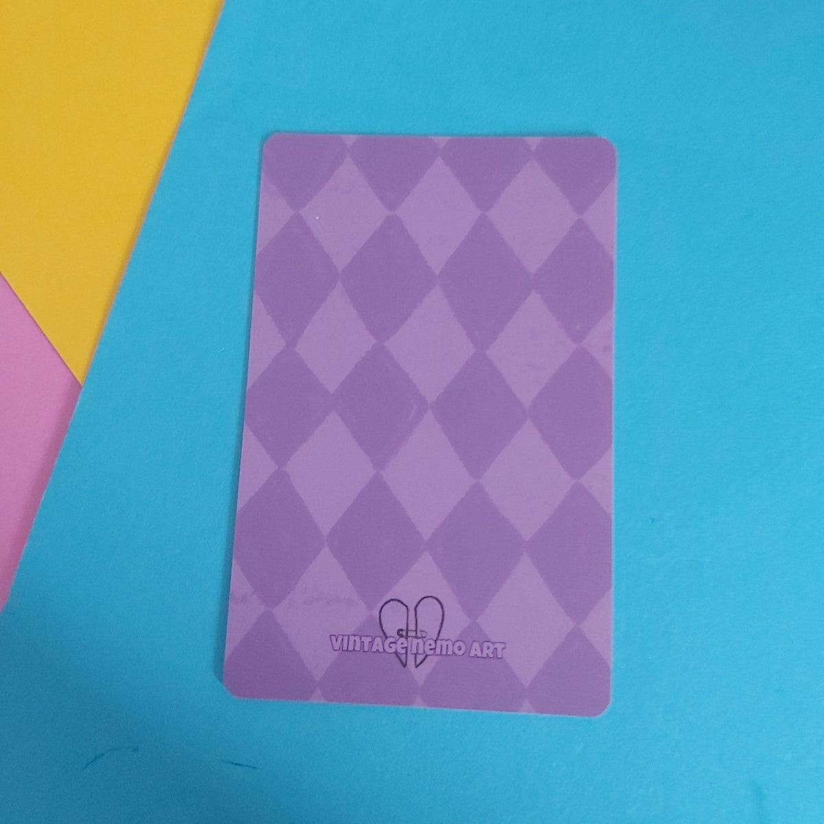 A rectangular purple card with a diamond harlequin pattern, featuring a small heart logo and the text 'VINTAGE NEMO ART' at the bottom, placed on a bright blue sheet of paper with yellow and pink paper visible in the corners.