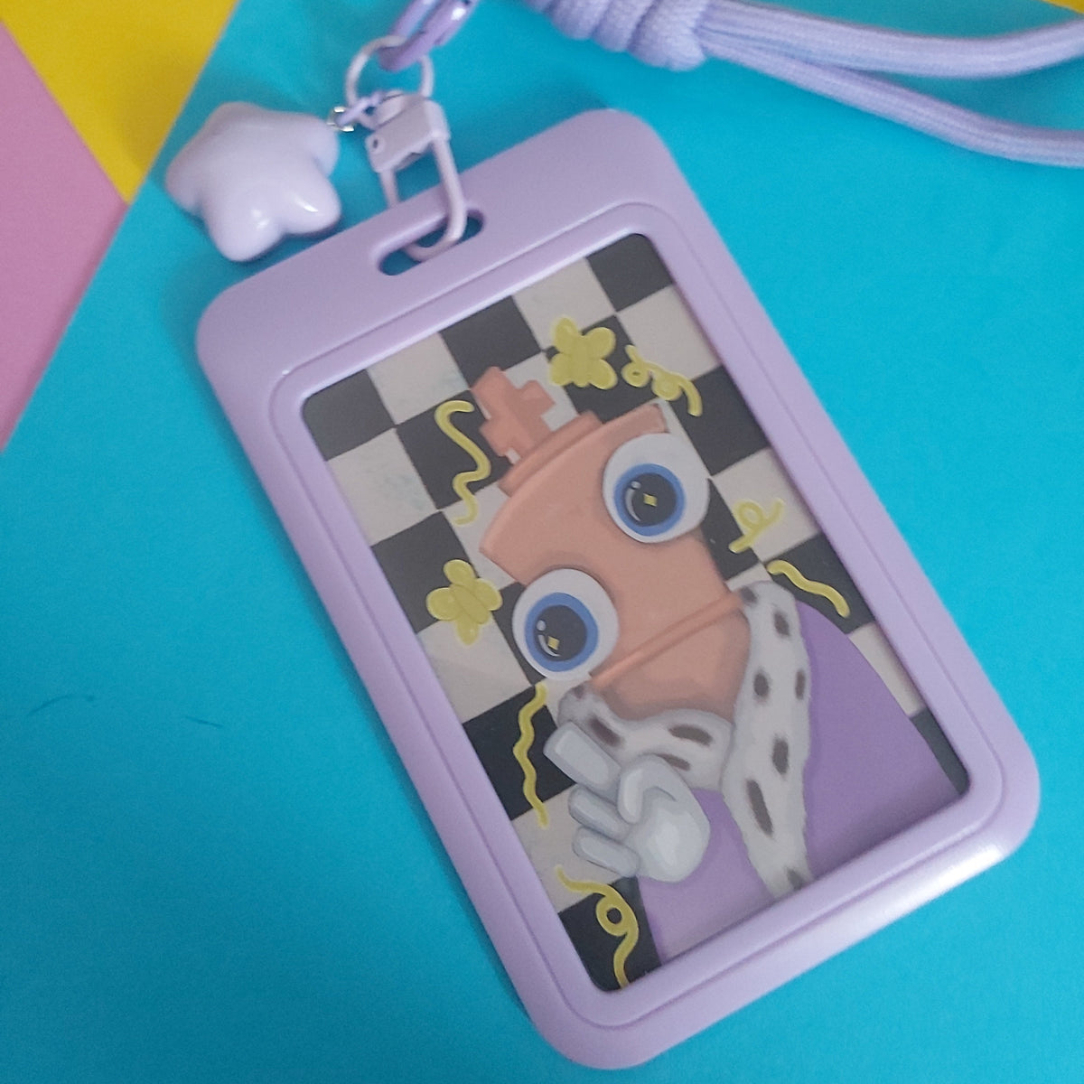 A lavender plastic ID badge holder with a matching braided lanyard and heart-shaped charm, featuring a colorful cartoon character printed on the front, displayed on a bright blue background.