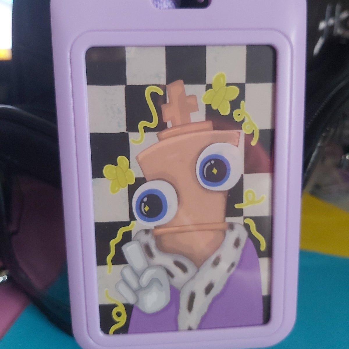 A lavender plastic ID badge holder featuring a custom printed graphic of a cartoonish character with a crown, blue eyes, and yellow flowers on a black-and-white checkered background.