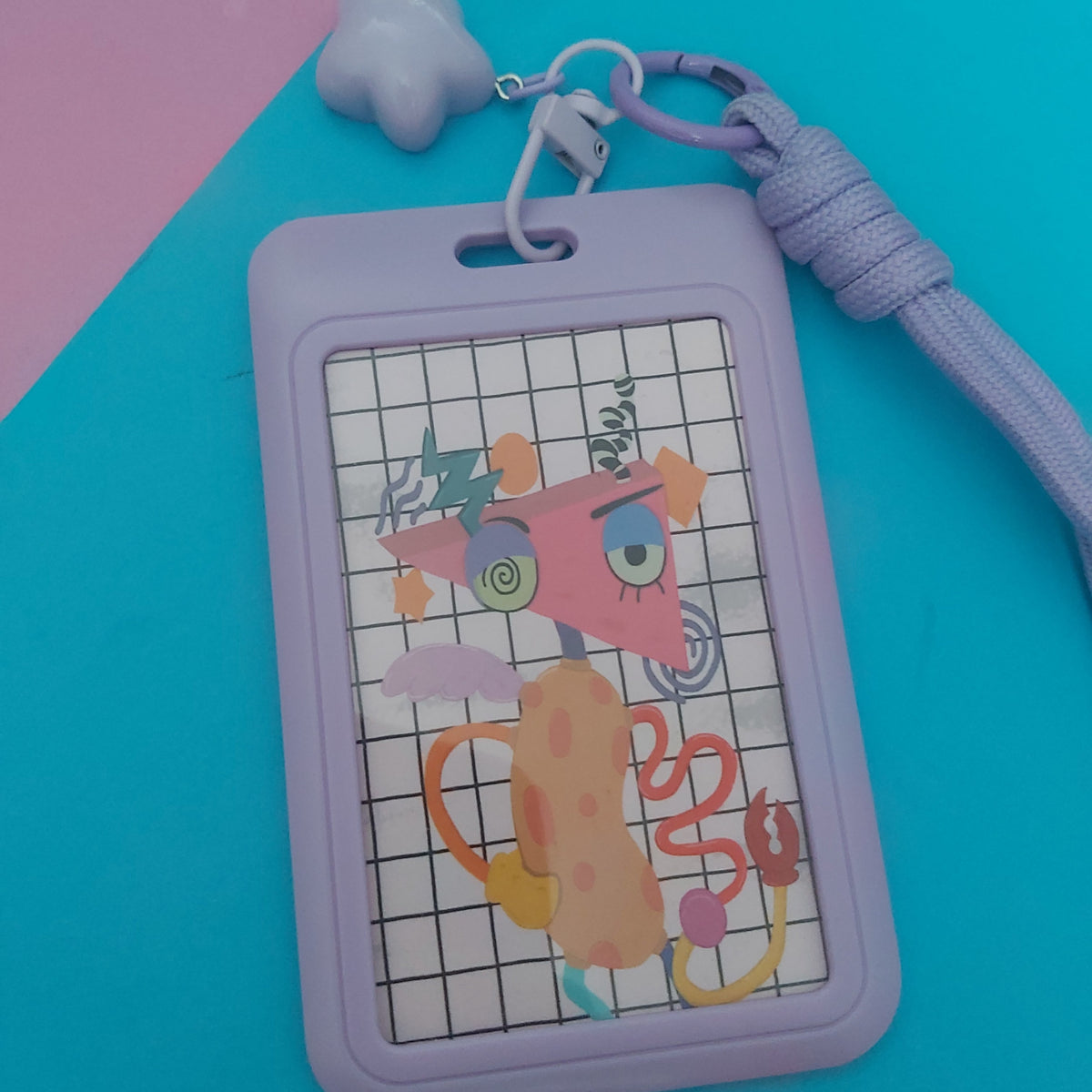 A lavender purple ID badge holder with a colorful abstract cartoon design on a grid background, featuring a matching purple wrist strap and star-shaped charm, set against a bright blue and pink background.