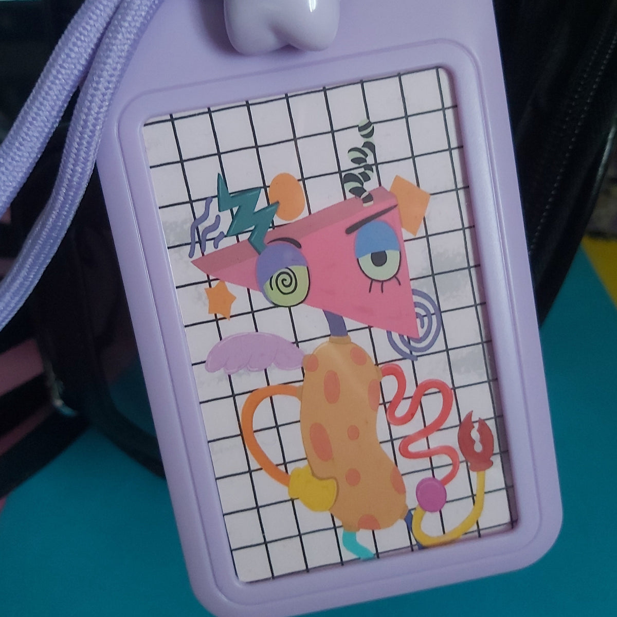 A lavender plastic ID badge holder with a matching braided wrist strap and a star-shaped charm, featuring a colorful abstract cartoon design on a white grid background.
