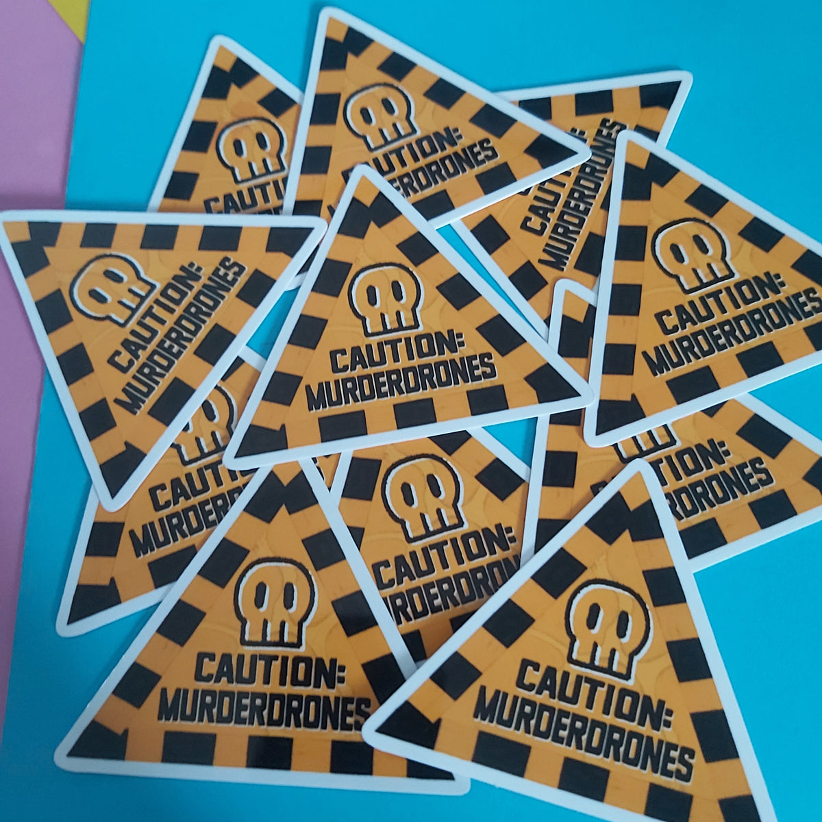 A pile of triangular stickers with a black and orange hazard pattern, featuring a white skull icon and the text 'CAUTION: MURDERDRONES' on a bright blue background.