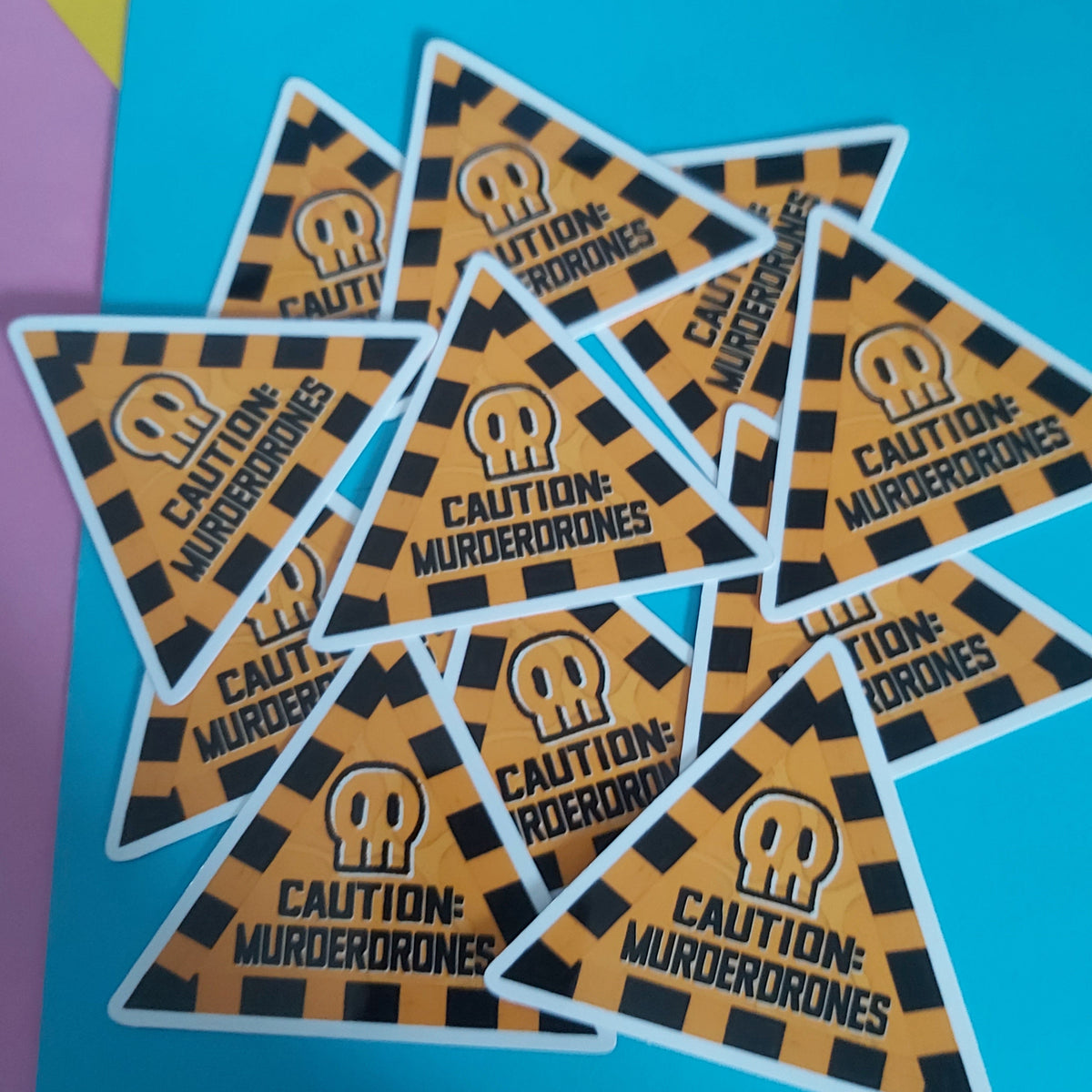 A group of triangular stickers with a black and orange hazard pattern, featuring a skull icon and the text 'CAUTION: MURDERDRONES' on a bright blue background.