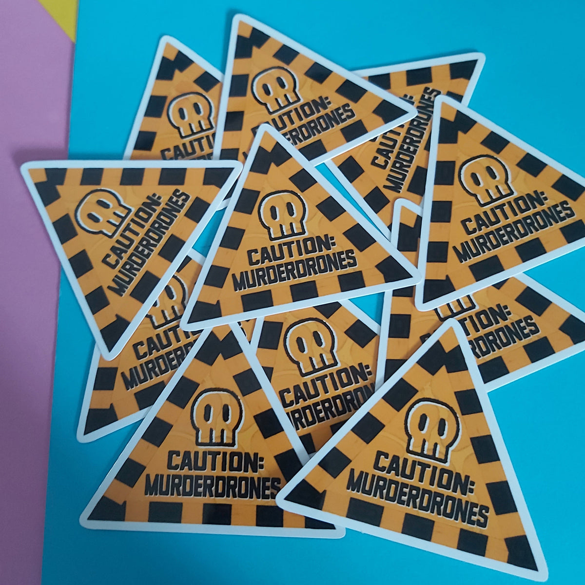 A pile of triangular stickers with a black and orange hazard stripe pattern, featuring a skull icon and the text 'CAUTION: MURDERDRONES' on a bright blue background.