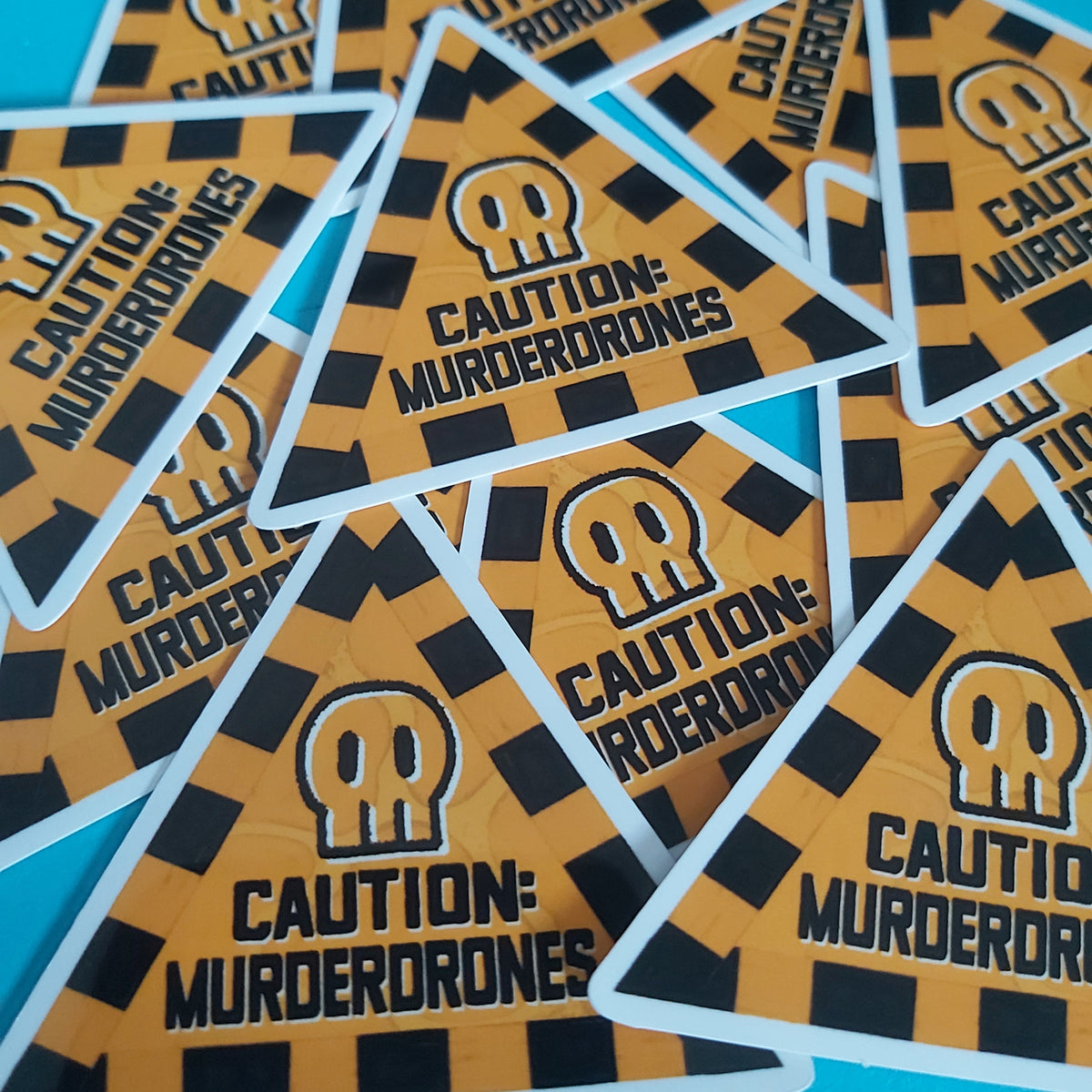 A pile of triangular warning stickers featuring a black and orange hazard stripe pattern, a white skull icon, and the text 'CAUTION: MURDERDRONES' on a bright blue background.