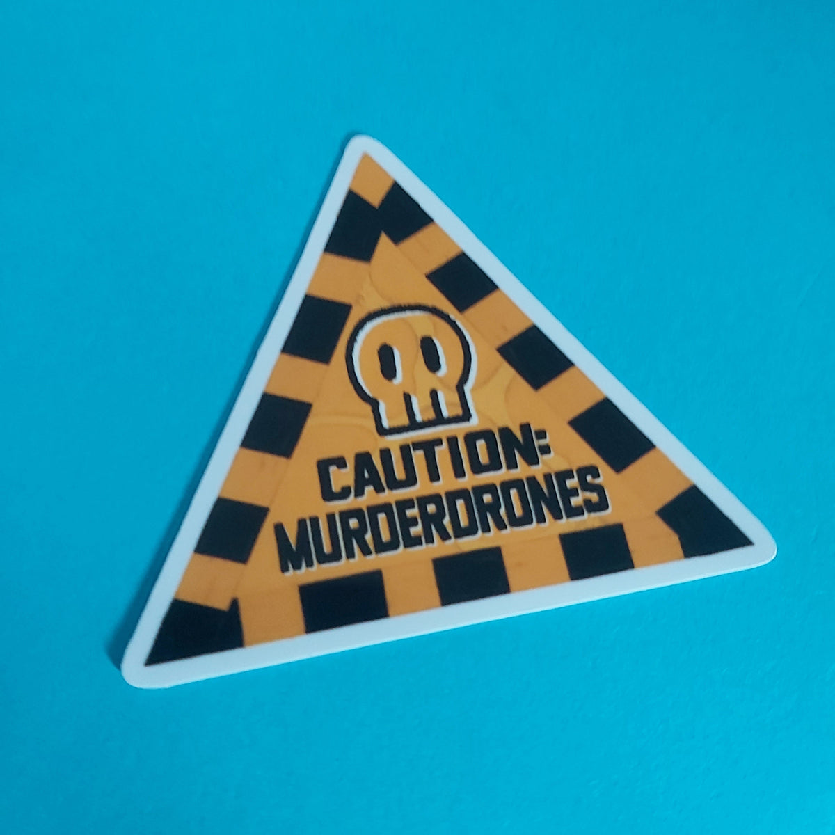 A triangular warning sticker with a yellow and black striped hazard pattern, a skull icon, and the text 'CAUTION: MURDERDRONES' on a bright blue background.