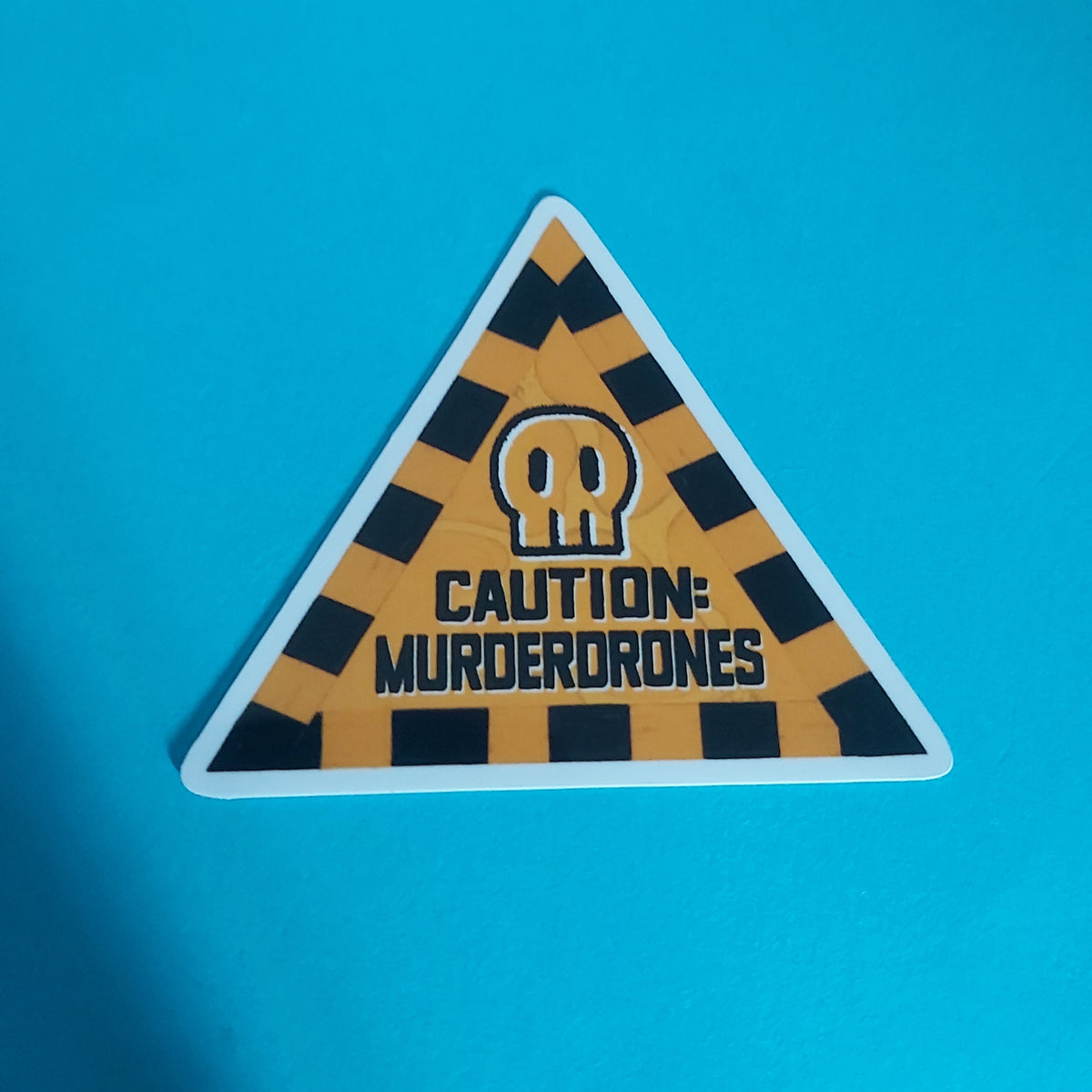 A triangular warning sticker with a yellow and black striped hazard pattern, a skull icon, and the text 'CAUTION: MURDERDRONES' on a bright blue background.