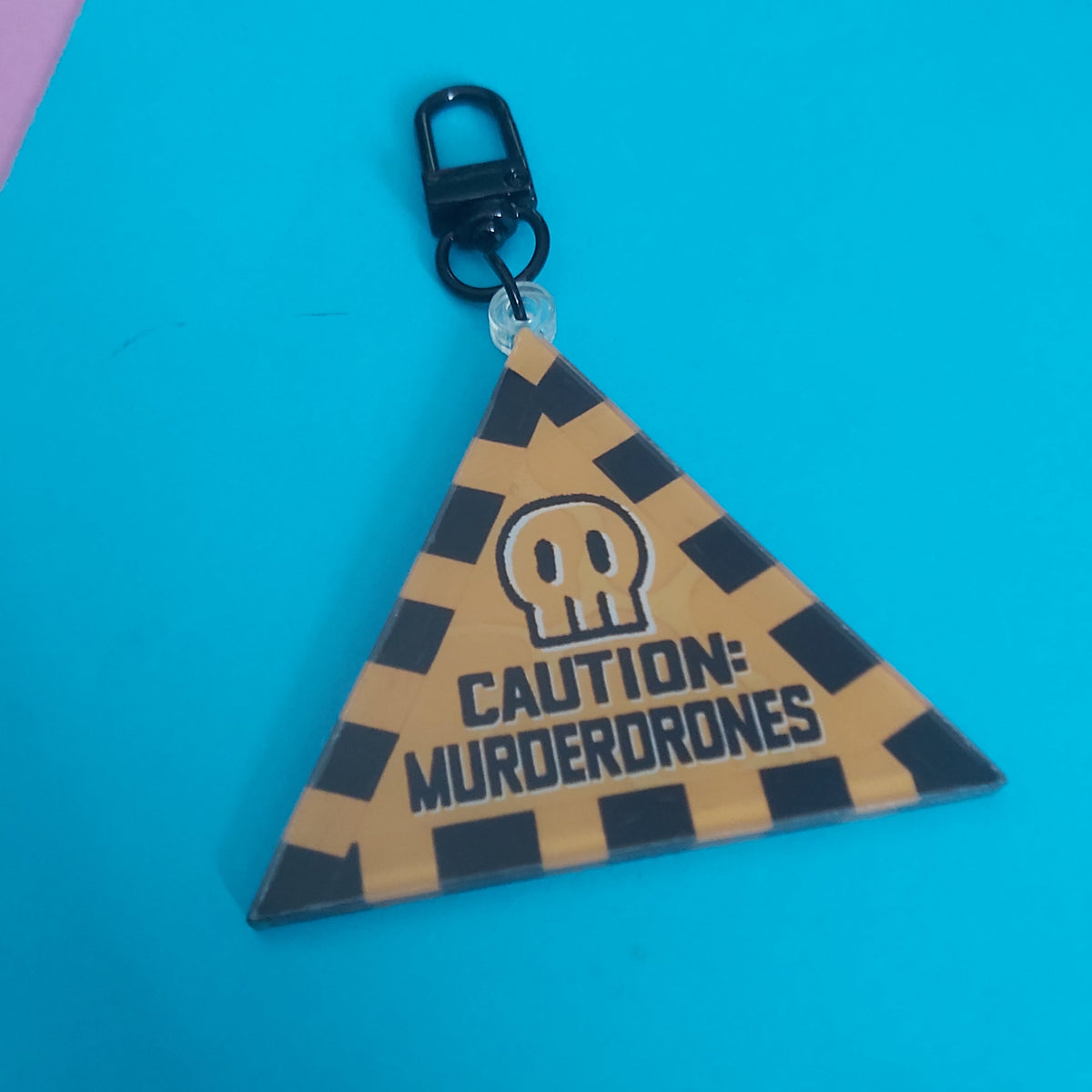 A triangular acrylic bag charm with a black and tan checkered pattern, featuring a skull icon and the text 'CAUTION: MURDERDRONES' on a bright blue background.
