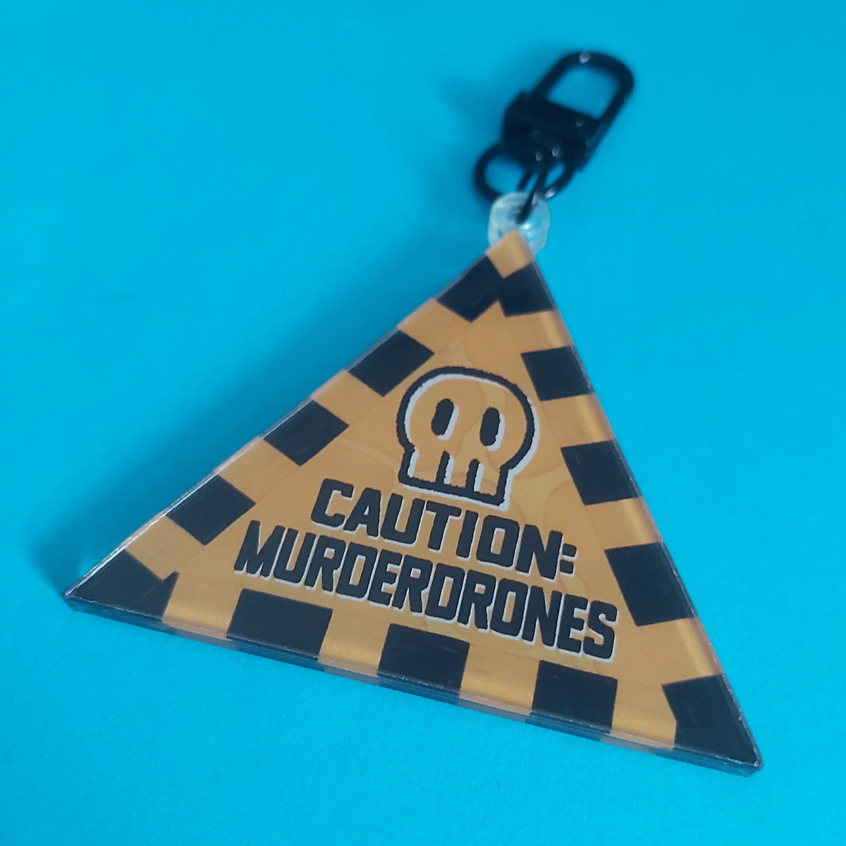 A triangular acrylic keychain featuring a black-and-yellow-orange striped caution pattern, a skull icon, and the text 'CAUTION: MURDERDRONES' in black, attached to a black metal clasp against a bright blue background.