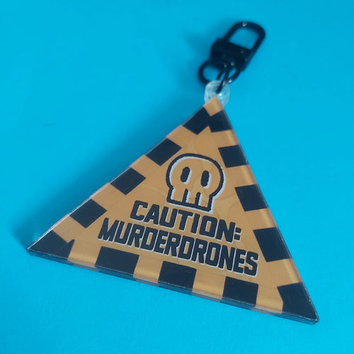 A triangular acrylic keychain featuring a black-and-yellow-orange striped caution pattern, a skull icon, and the text 'CAUTION: MURDERDRONES' in black, attached to a black metal clasp against a bright blue background.
