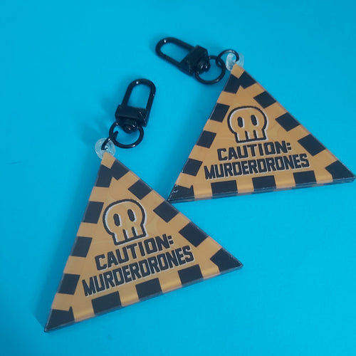 Two triangular acrylic keychains with a black and yellow-orange caution-stripe pattern, featuring a skull icon and the text 'CAUTION: MURDERDRONES', attached to black swivel clasps, displayed on a bright blue background.