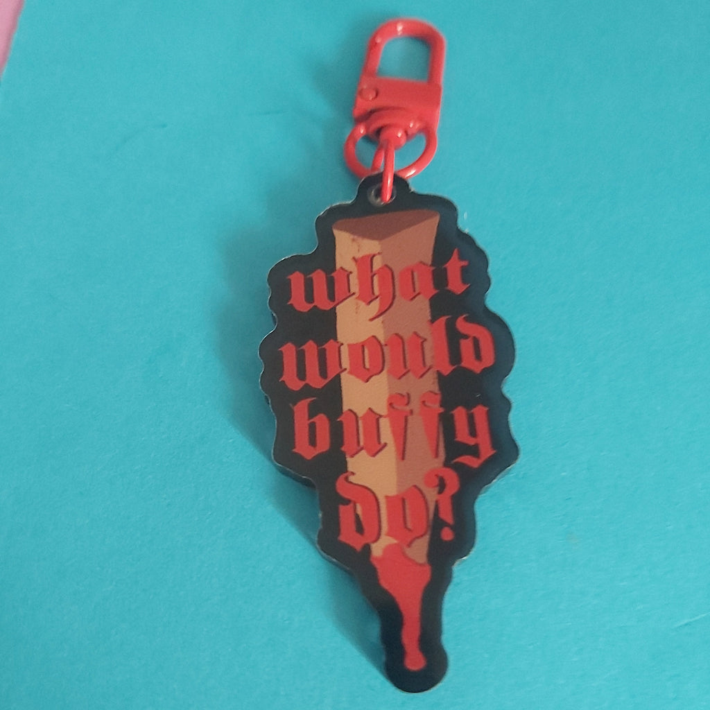 A bag charm shaped like a stake, featuring the gothic text 'what would buffy do?' in red on a black and brown gradient background, with a red metal clasp at the top, set against a bright turquoise background.