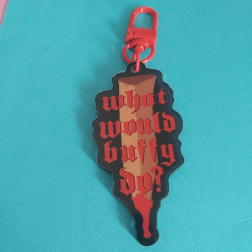 A bag charm shaped like a stake, featuring the gothic text 'what would buffy do?' in red on a black and brown gradient background, with a red metal clasp at the top, set against a bright turquoise background.