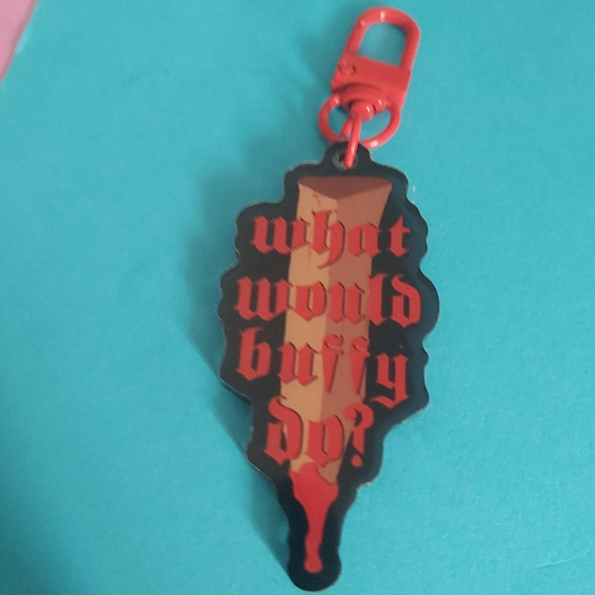 A keychain bag charm shaped like a stake, featuring the text 'what would buffy do?' in red gothic lettering. The stake has a black outline, a brown handle, and is attached to a red metal clasp.