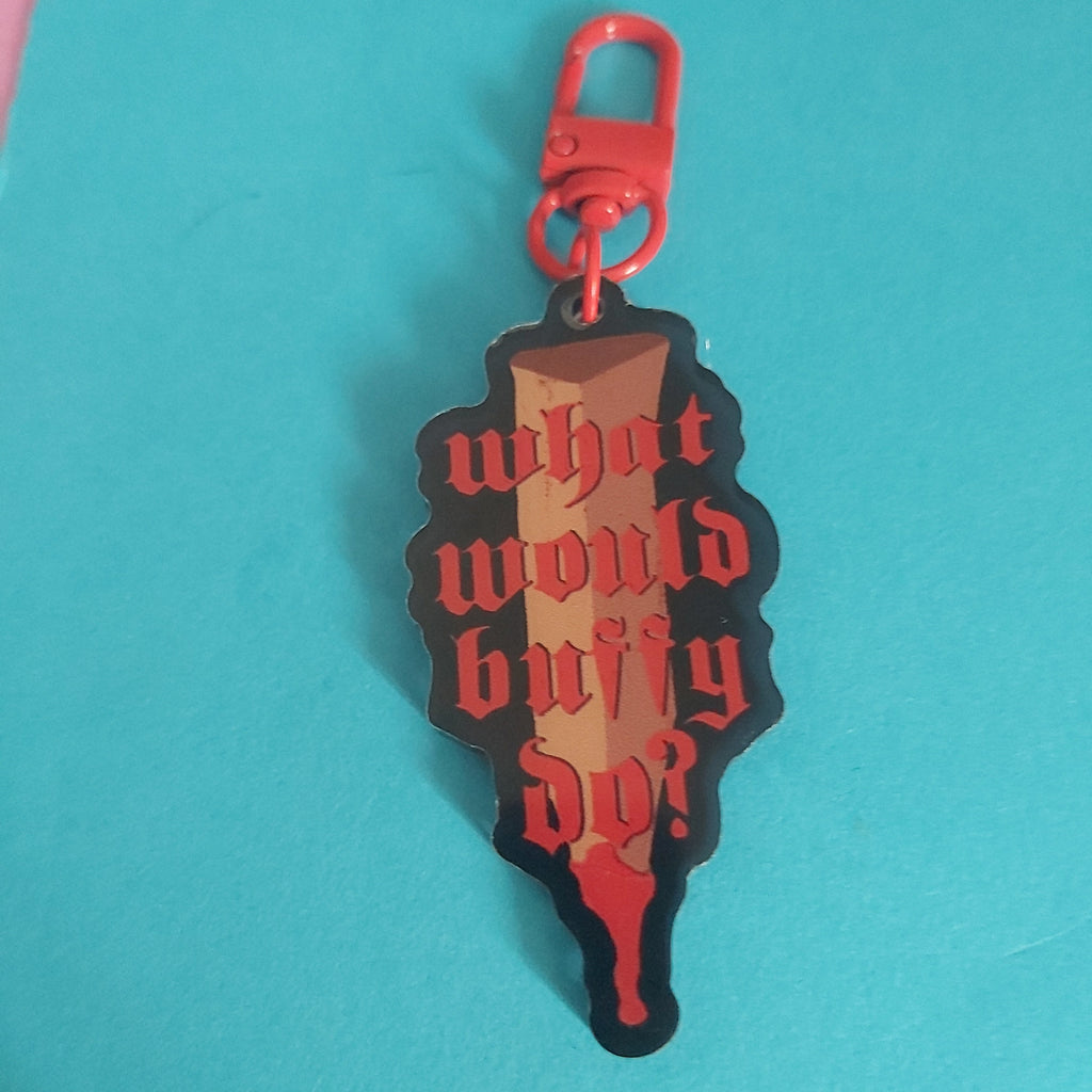 A die-cut acrylic keychain charm featuring a stake design with the gothic text 'what would buffy do?' in red, set against a black and brown background, attached to a red lobster clasp. The keychain is photographed on a bright turquoise surface.