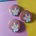 Three round pinback buttons featuring a cartoon character with red hair and a button eye, arranged on a split pink and yellow background with a blue strip at the top.