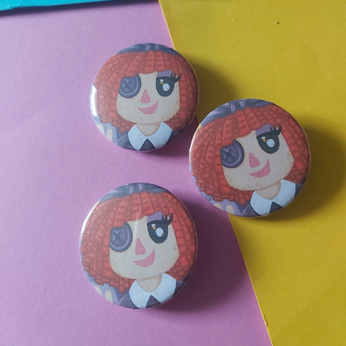 Three round pinback buttons featuring a cartoon character with red hair and a button eye, arranged on a split pink and yellow background with a blue strip at the top.